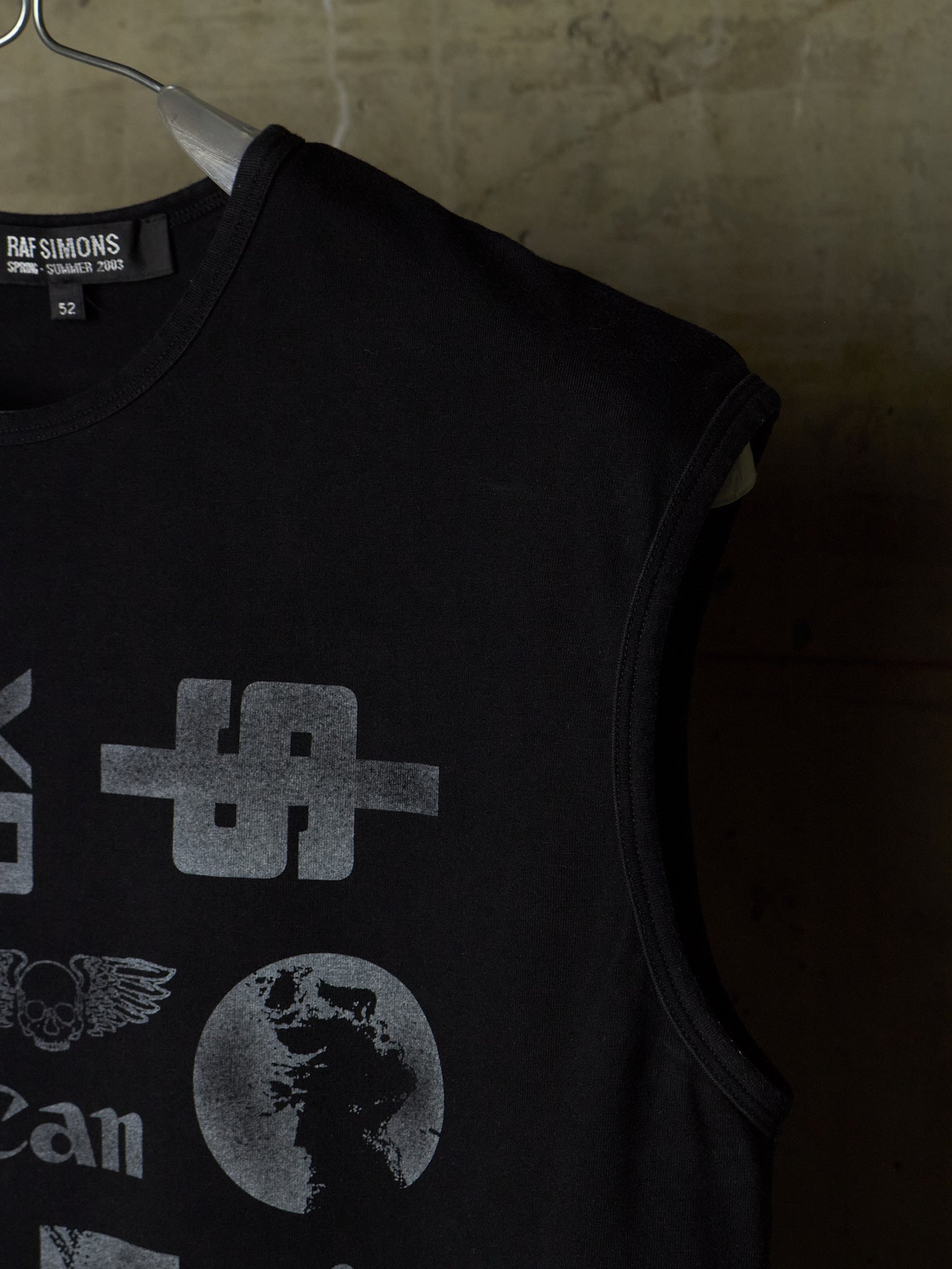 A black sleeveless tank top hanging on a metal hanger against a textured concrete wall. The shirt features a distressed gray graphic print with a stylized symbol, a winged emblem, and a globe design.
