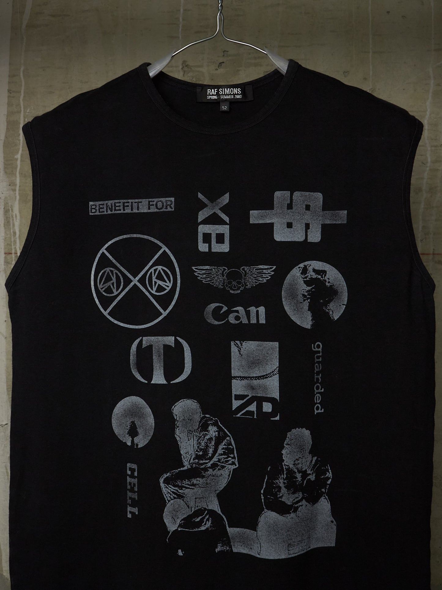 A black sleeveless t-shirt hanging on a metal hanger against a textured concrete wall. The shirt features a distressed, faded gray graphic print of various punk and alternative music band logos, including X-Bits, Can, Cradle of Filth, and Cell.
