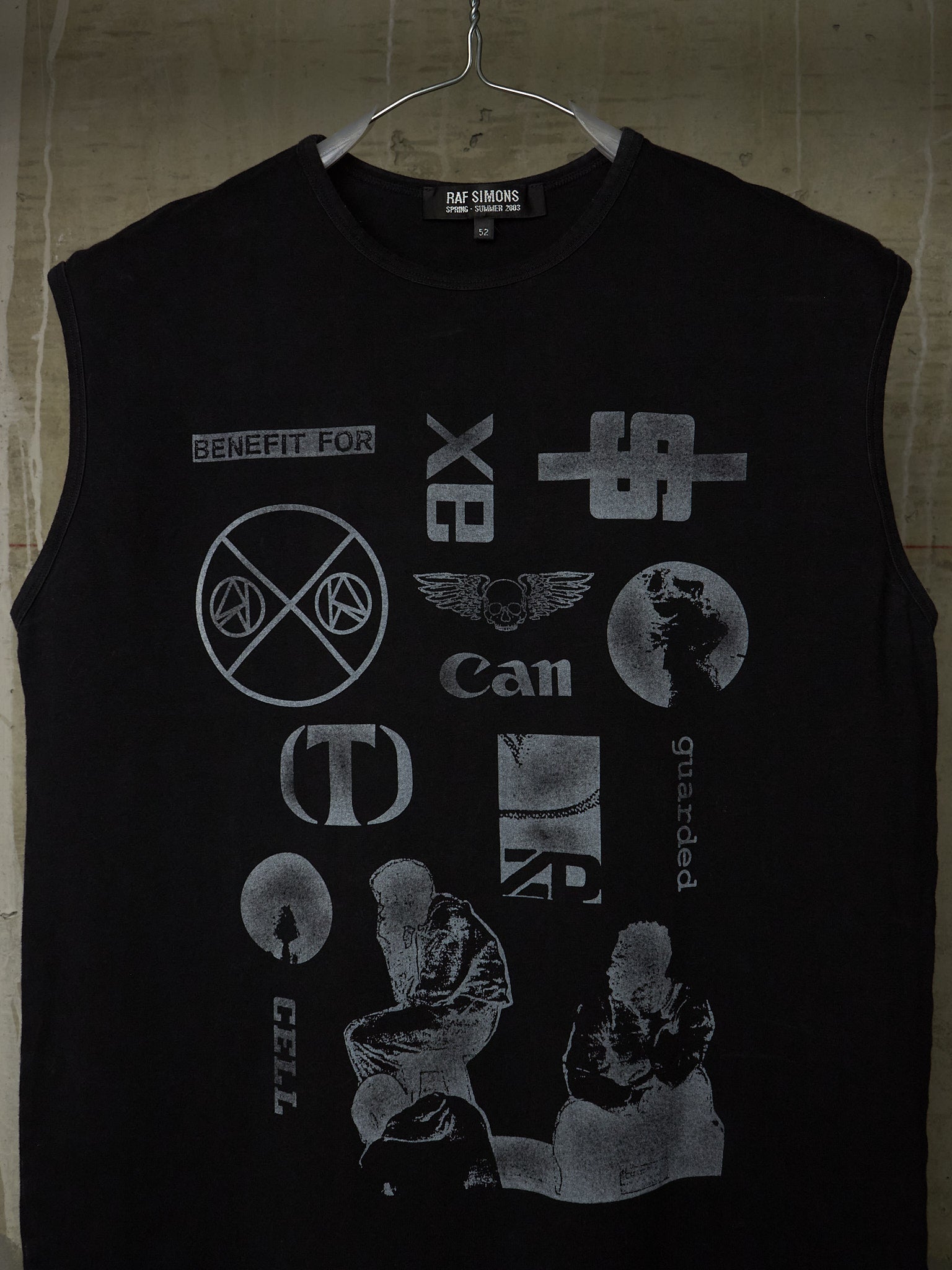 A black sleeveless t-shirt hanging on a metal hanger against a textured concrete wall. The shirt features a distressed, faded gray graphic print of various punk and alternative music band logos, including X-Bits, Can, Cradle of Filth, and Cell.