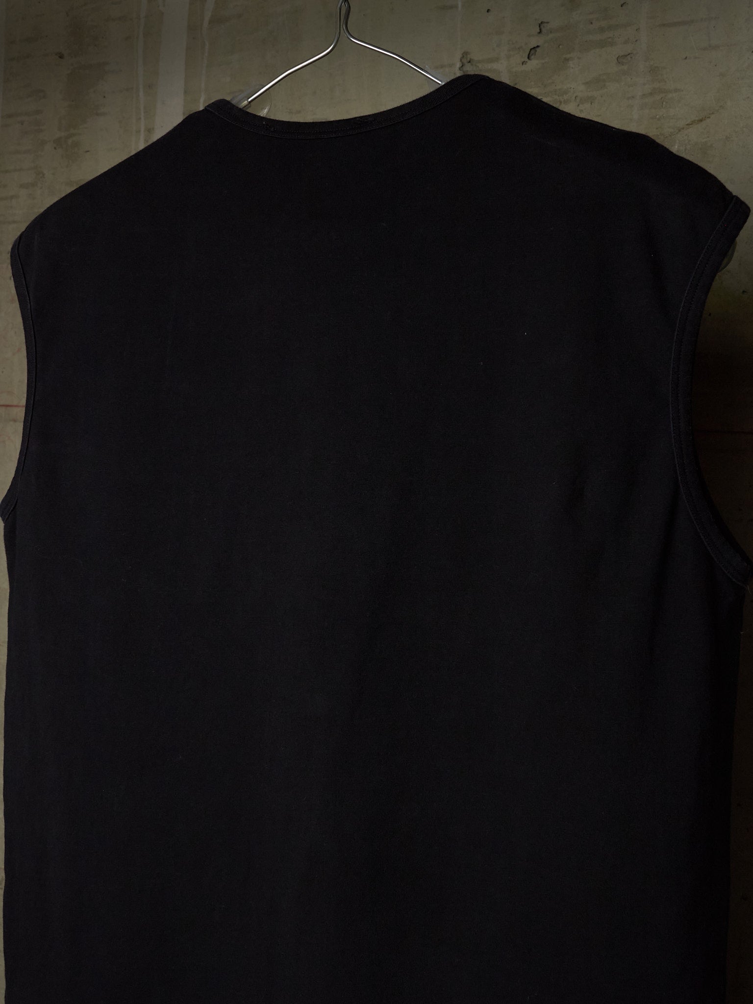 A black sleeveless ribbed tank top hanging on a metal hanger against a textured gray wall.