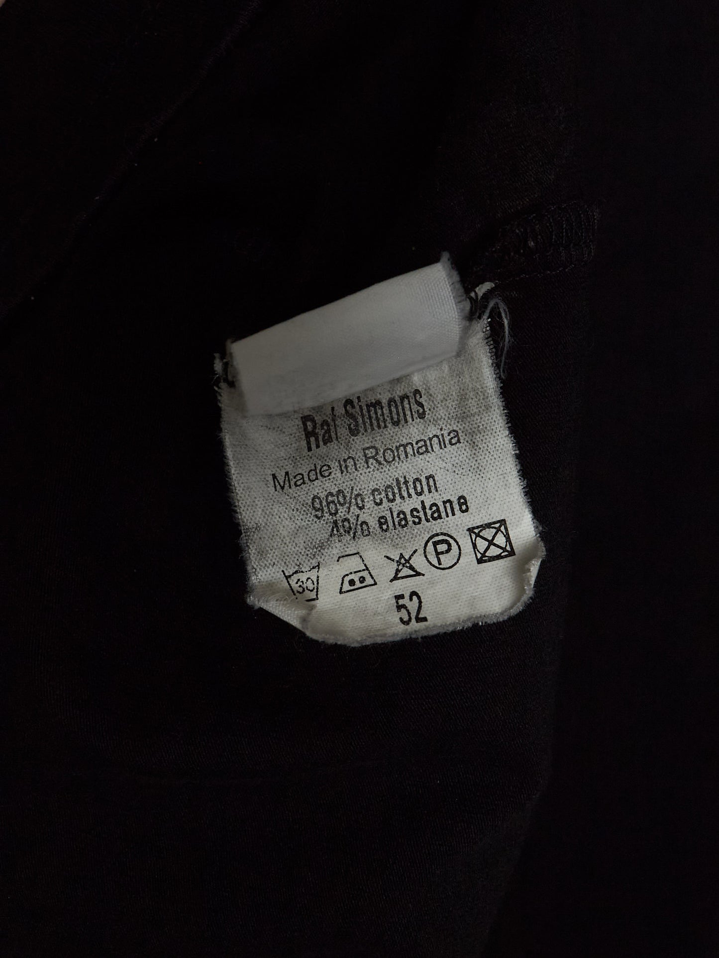 A close-up of a white fabric care label sewn onto a black garment. The label reads 'Rai Simons' and shows care instructions and the size number 52.
