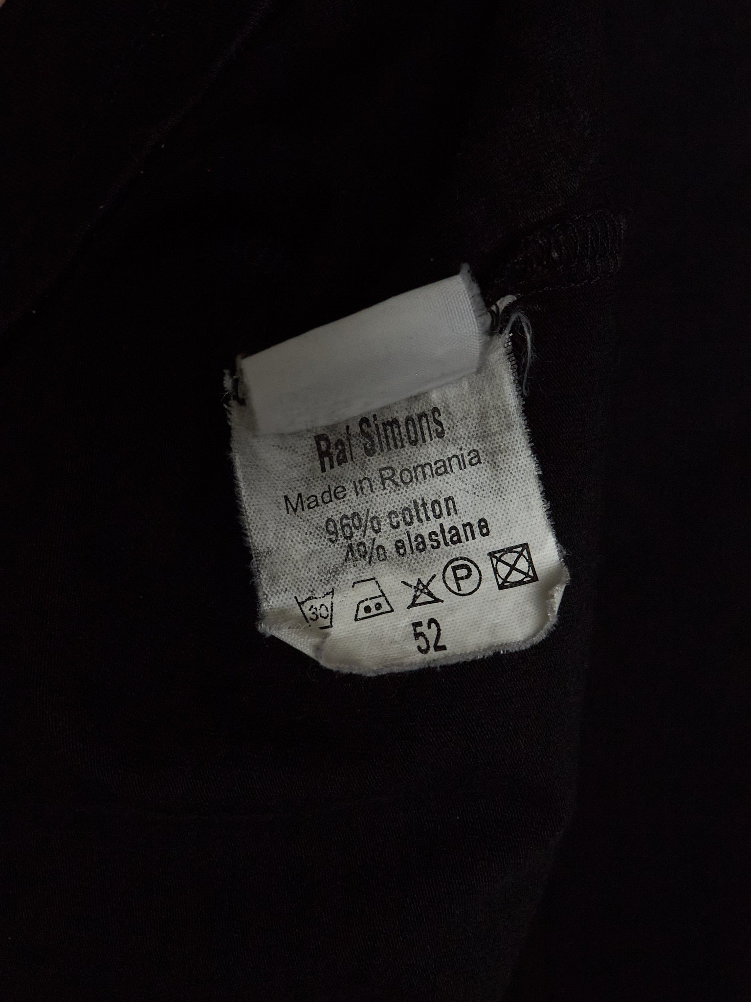 A close-up of a white fabric care label sewn onto a black garment. The label reads 'Rai Simons' and shows care instructions and the size number 52.