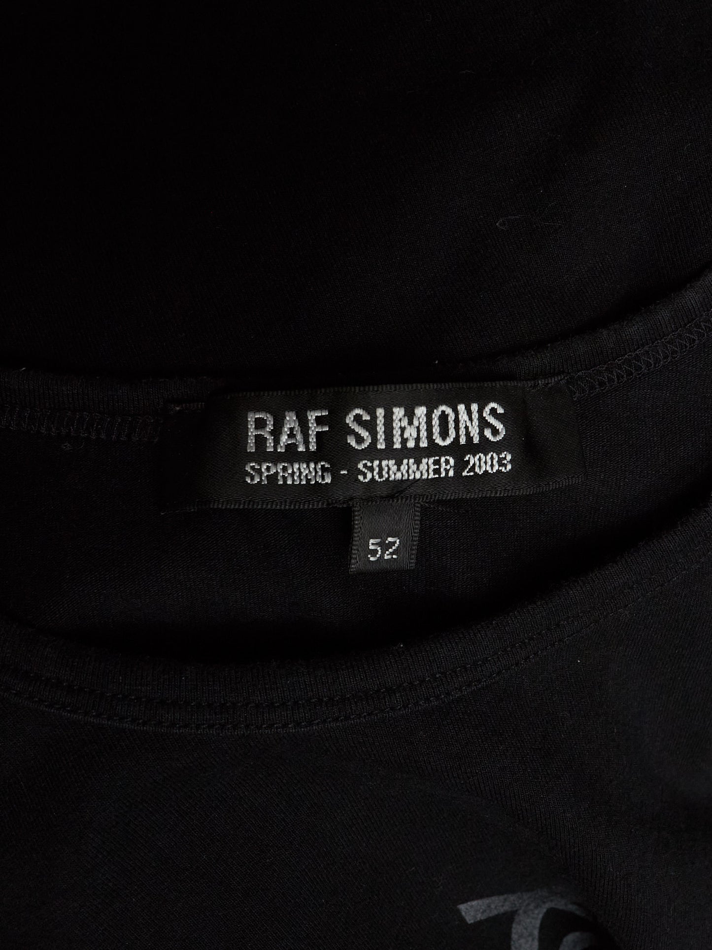 Close-up of a black Raf Simons t-shirt neck label reading 'RAF SIMONS SPRING - SUMMER 2003' and size '52'.