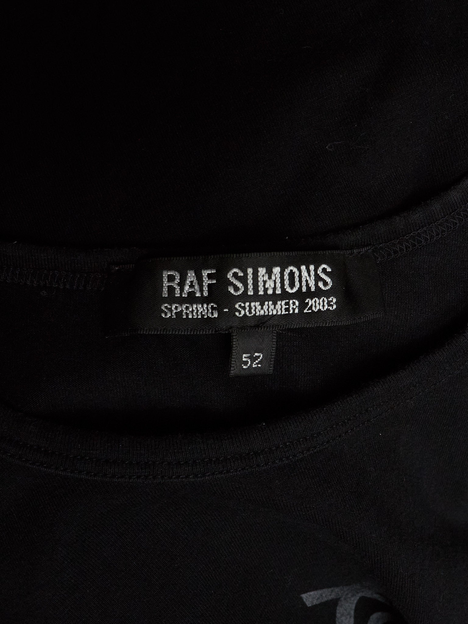 Close-up of a black Raf Simons t-shirt neck label reading 'RAF SIMONS SPRING - SUMMER 2003' and size '52'.