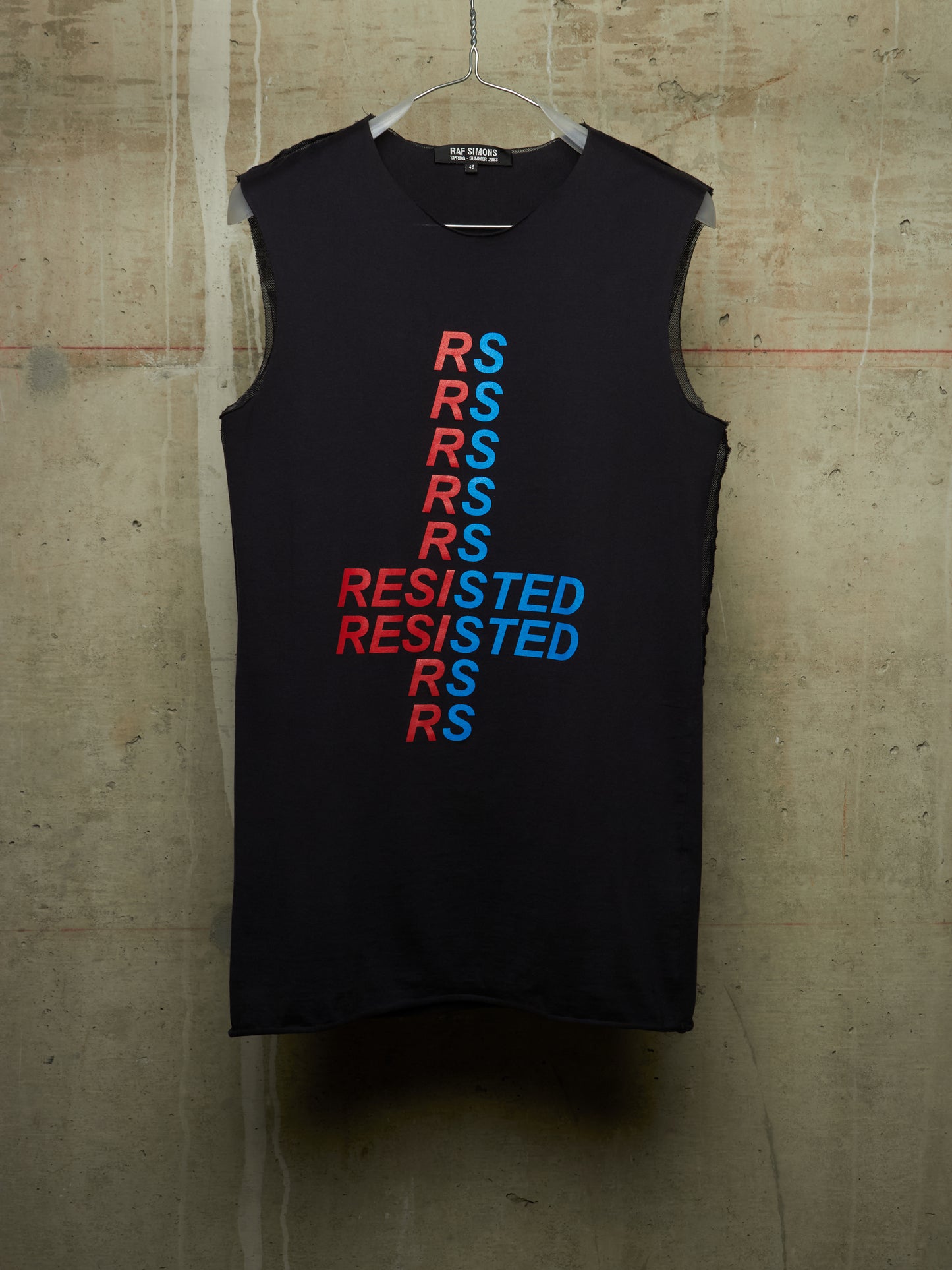 A sleeveless black tank top hanging on a wire hanger against a textured concrete wall. The front features a vertical graphic design spelling 'RESISTED' in alternating red and blue text.