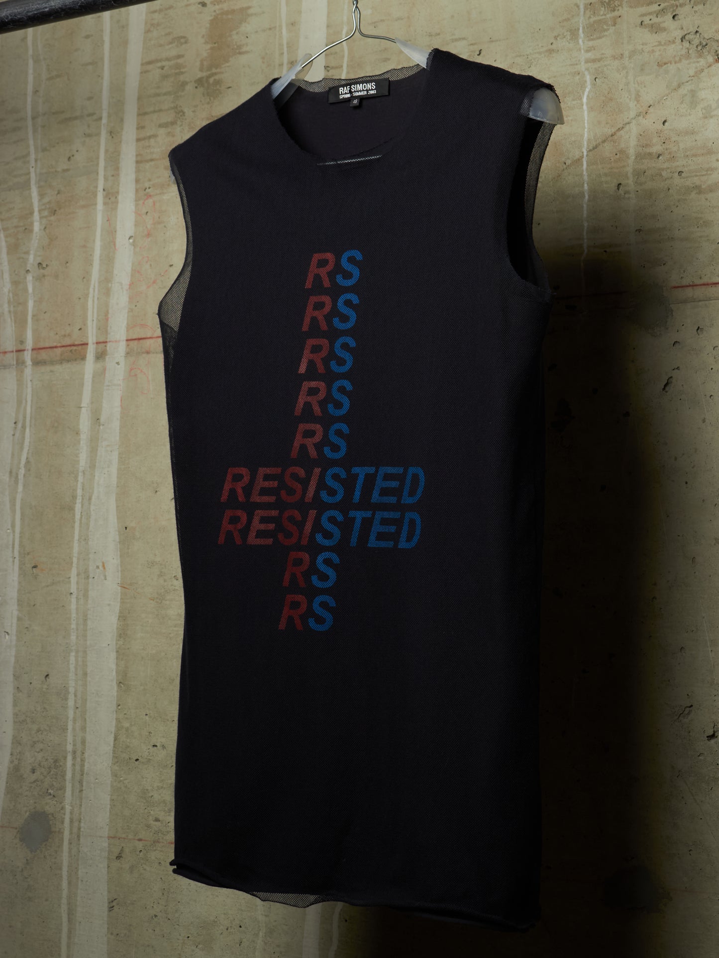 A sleeveless black tank top hanging on a wire hanger against a textured concrete wall. The front features a vertical graphic design with the letters 'RS' repeated in red and blue, spelling out 'RESISTED' vertically.