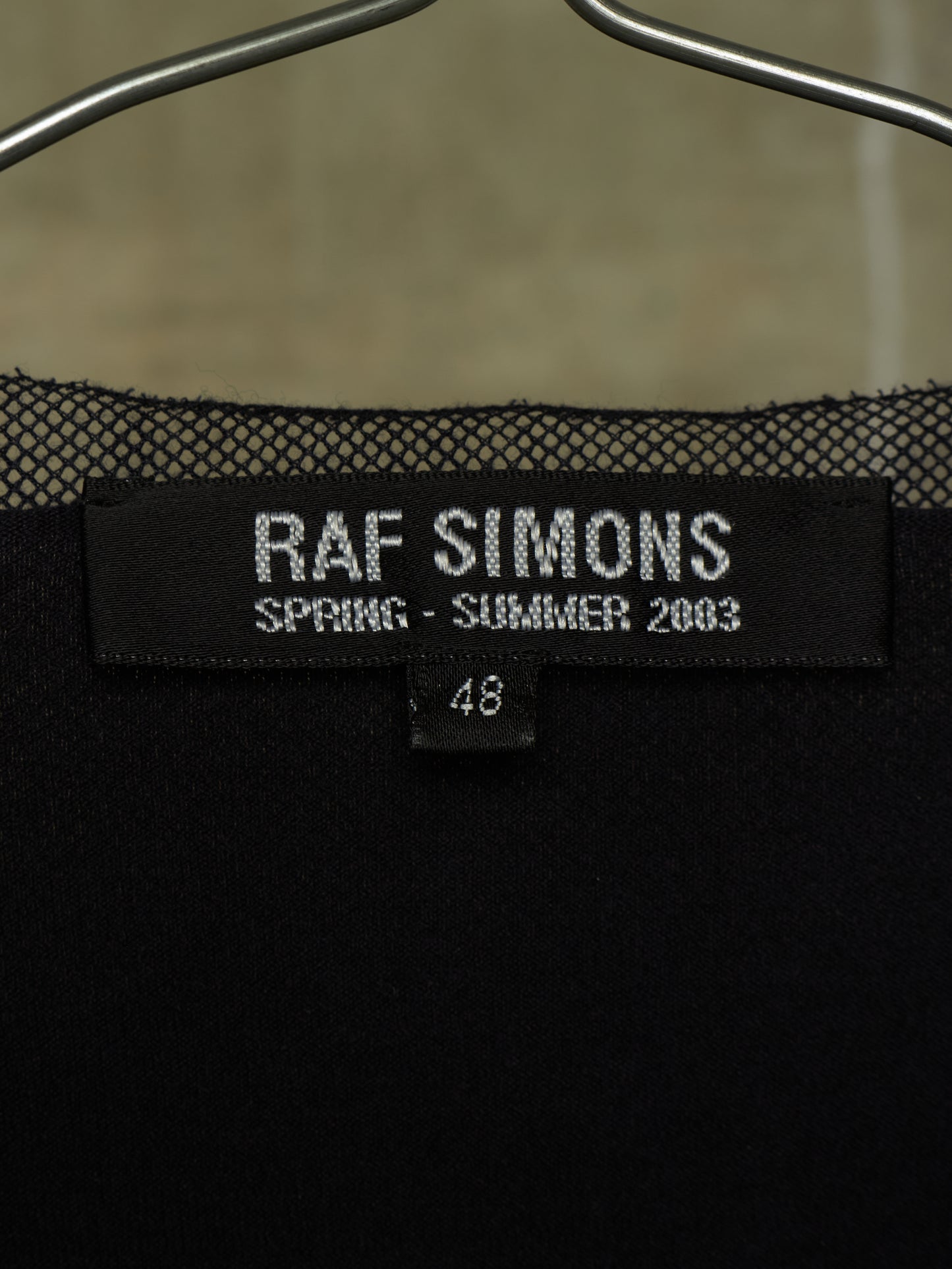 Close-up of a black Raf Simons Spring-Summer 2003 knit garment on a hanger, showing the brand label and size 48.