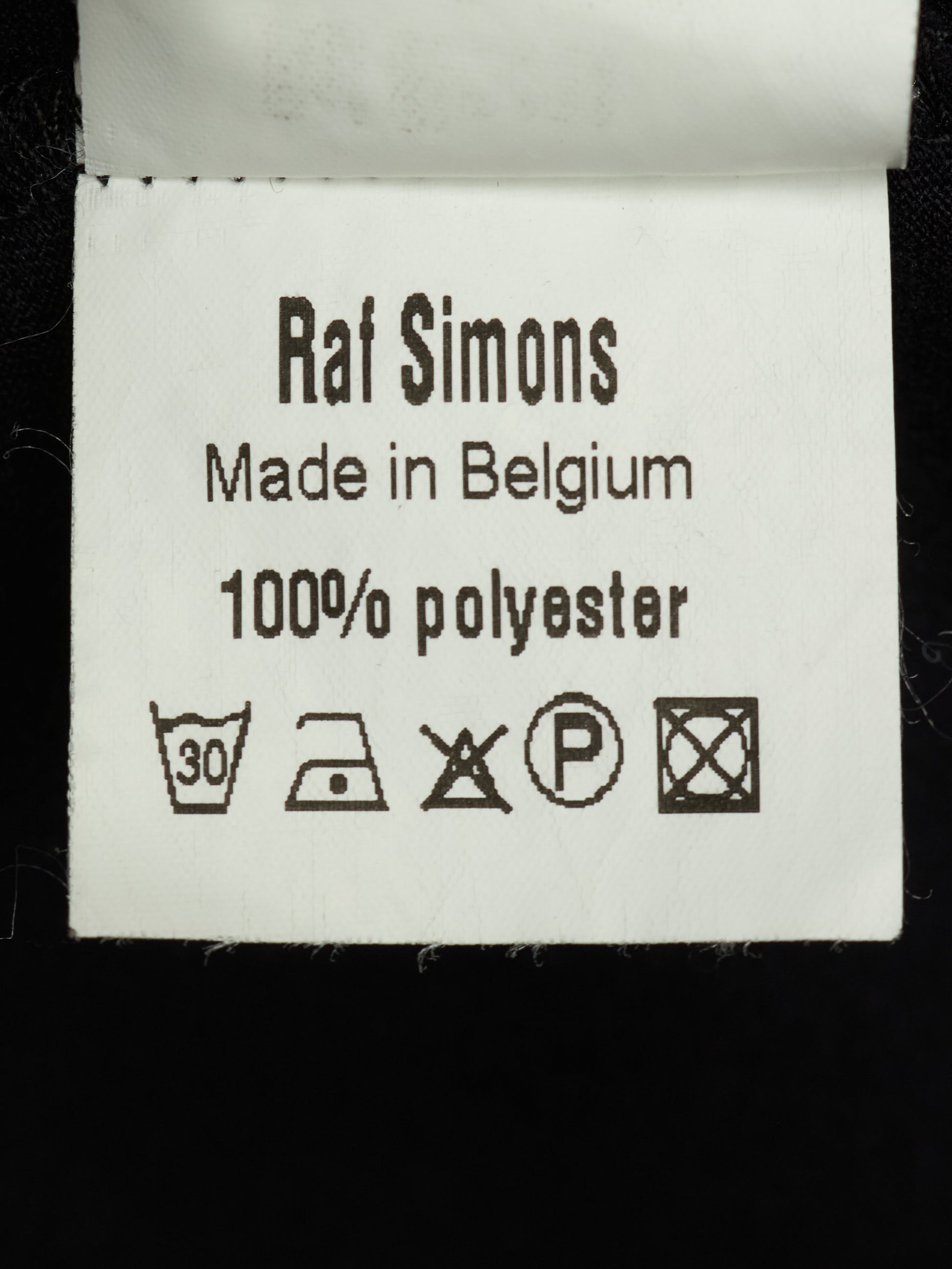 A close-up of a white fabric care label reading 'Raf Simons Made in Belgium 100% polyester' with standard washing instruction symbols below, set against a black background.