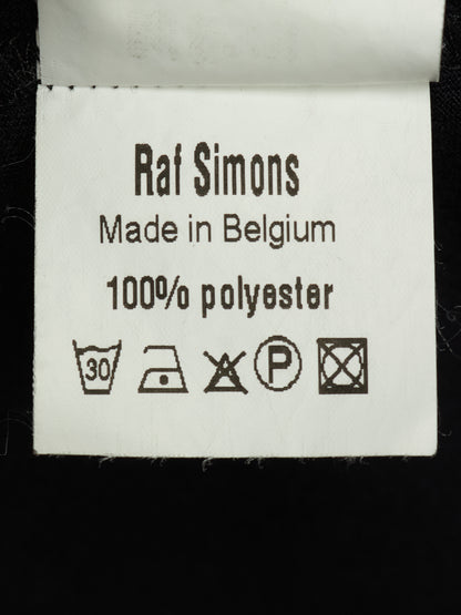 A close-up of a white fabric care label reading 'Raf Simons Made in Belgium 100% polyester' with standard washing instruction symbols below, set against a black background.