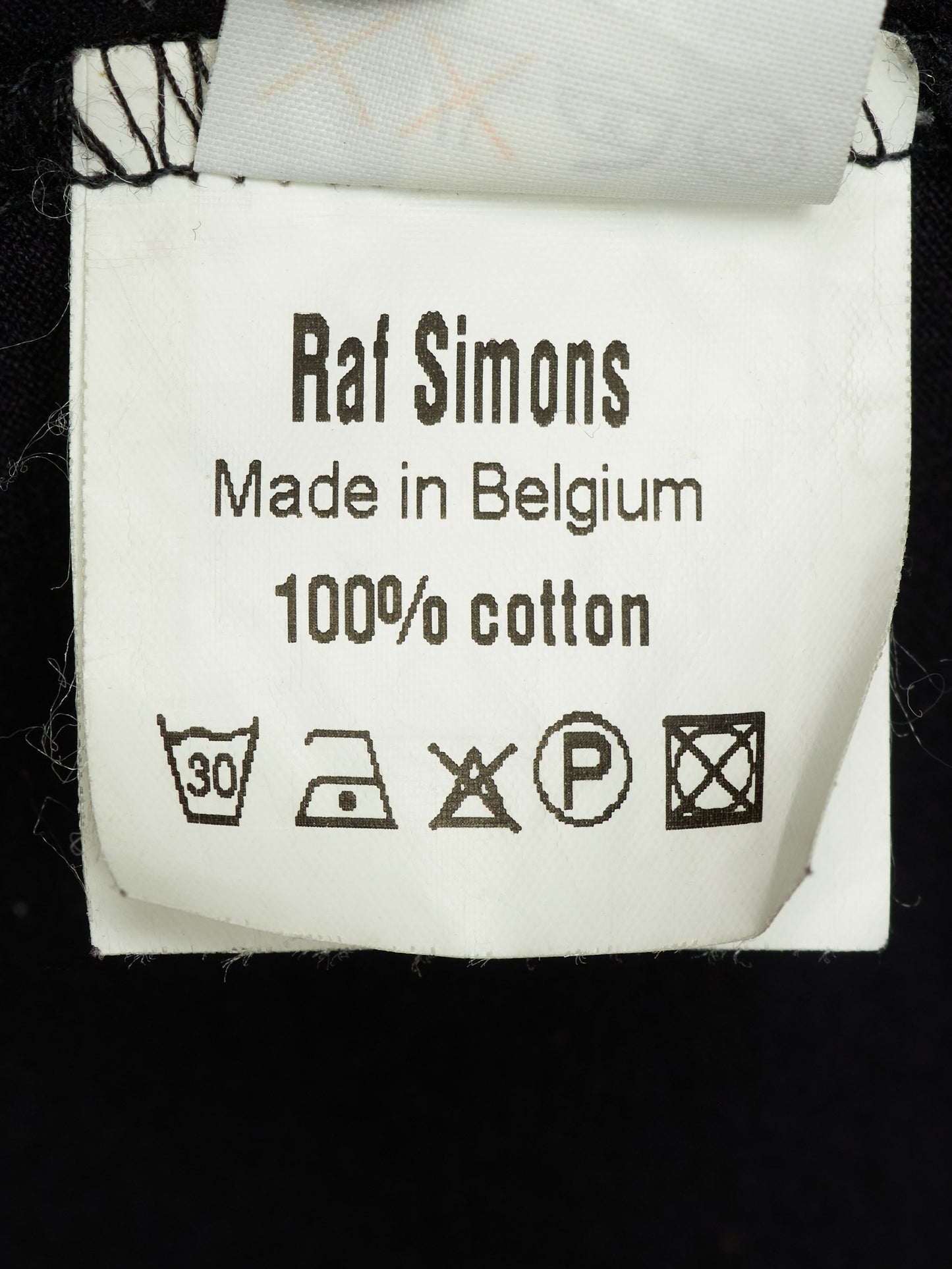A close-up of a white cotton garment label reading 'Raf Simons Made in Belgium 100% cotton' with care symbols, attached to a black garment.