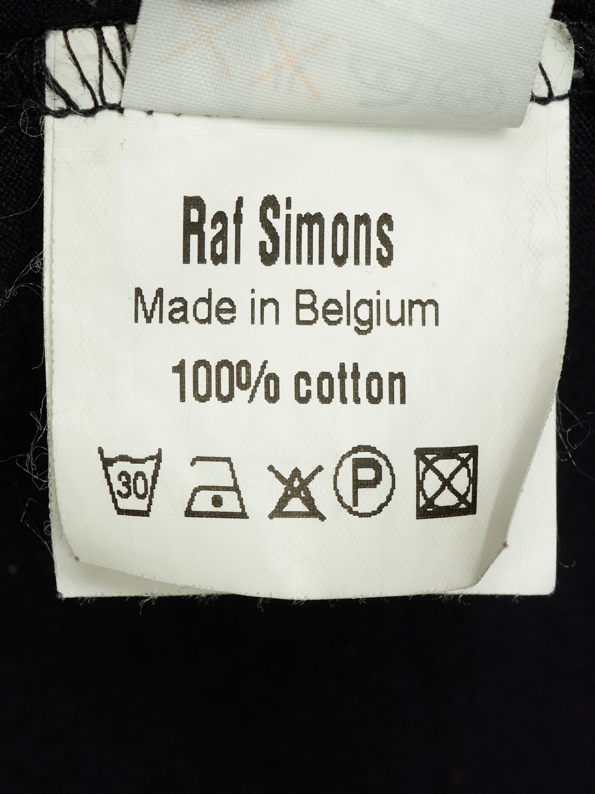 A close-up of a white cotton garment label reading 'Raf Simons Made in Belgium 100% cotton' with care symbols, attached to a black garment.