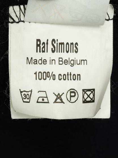 A close-up of a white cotton garment label reading 'Raf Simons Made in Belgium 100% cotton' with care symbols, attached to a black garment.
