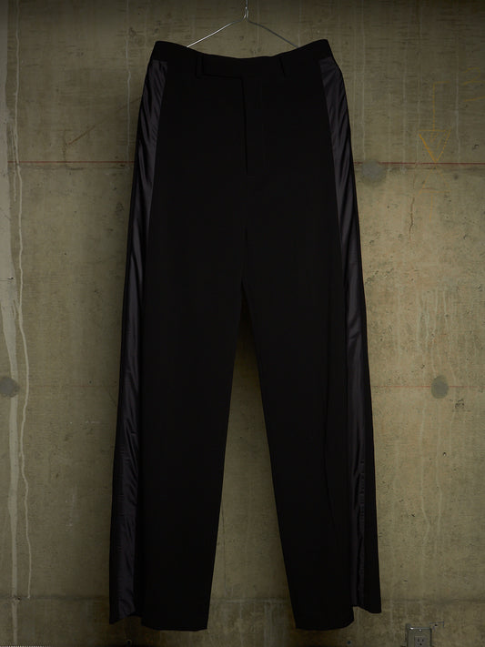 Black wide-leg trousers with dark satin side panels, hanging on a wire hanger against a textured concrete wall.