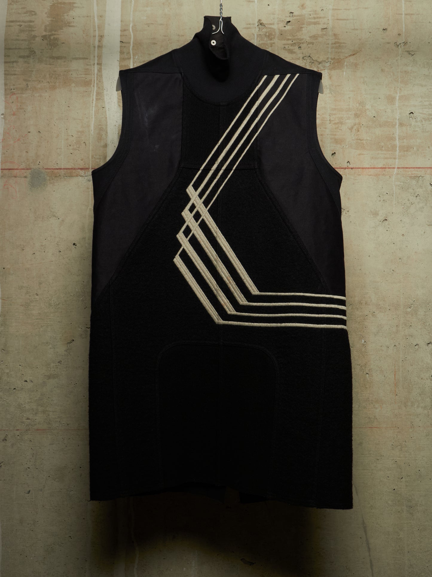A sleeveless black dress with a mock neck and geometric beige stripe patterns on the front, hanging against a textured concrete wall.