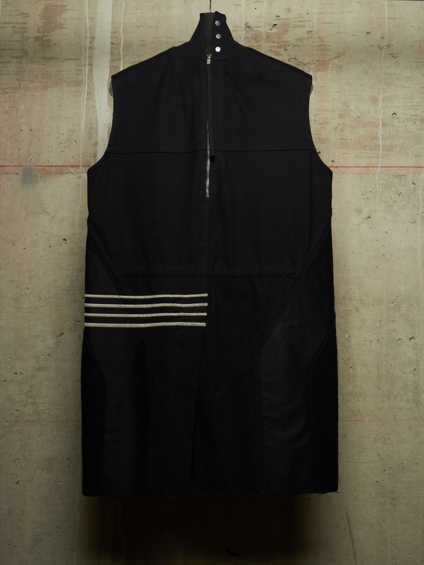 A black sleeveless zip-front vest or longline top hanging against a textured concrete wall, featuring horizontal beige stripes near the lower left side.