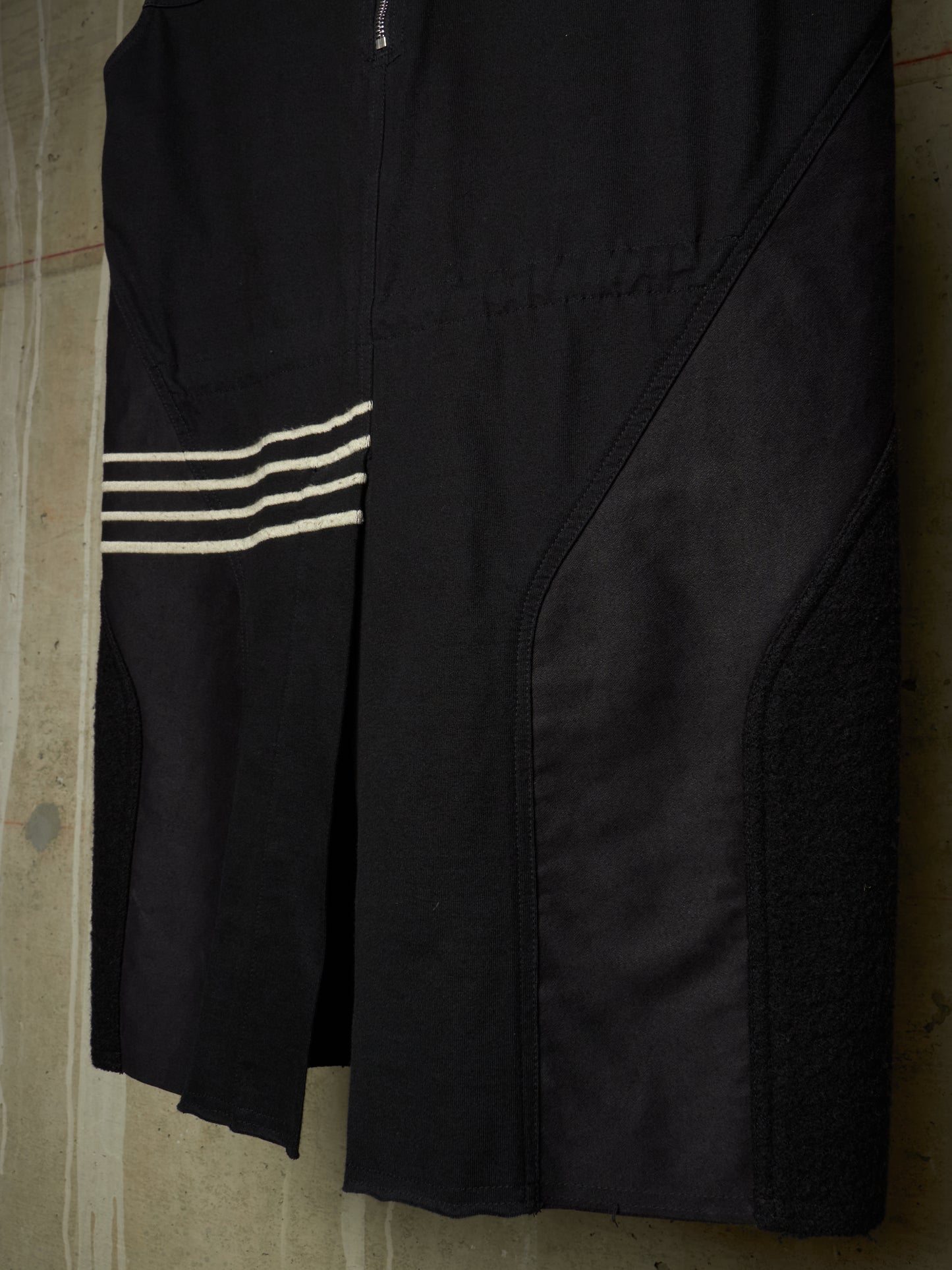 A close-up of a black athletic garment, likely a cycling shorts or bib shorts, featuring three horizontal white stripes on the left side and a zippered front closure, shown hanging against a concrete wall.