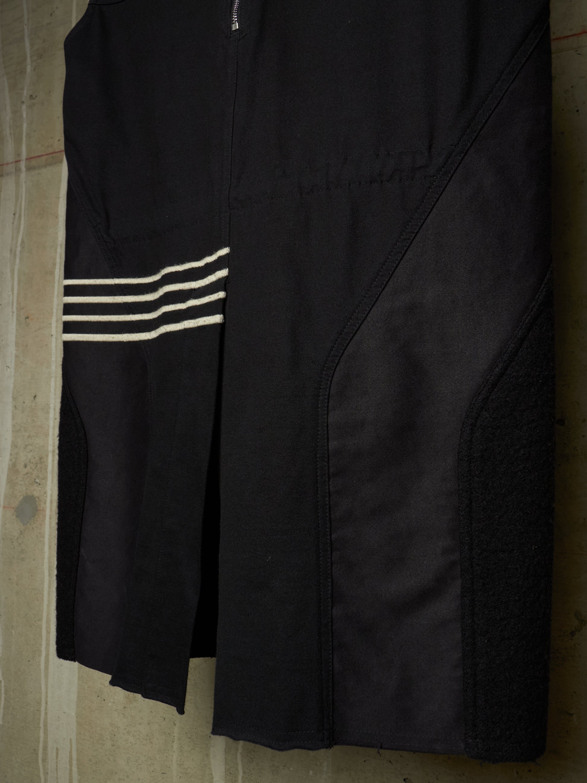 A close-up of a black athletic garment, likely a cycling shorts or bib shorts, featuring three horizontal white stripes on the left side and a zippered front closure, shown hanging against a concrete wall.