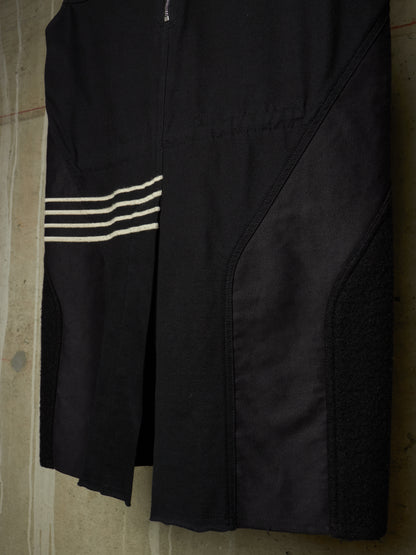 A close-up of a black athletic garment, likely a cycling shorts or bib shorts, featuring three horizontal white stripes on the left side and a zippered front closure, shown hanging against a concrete wall.