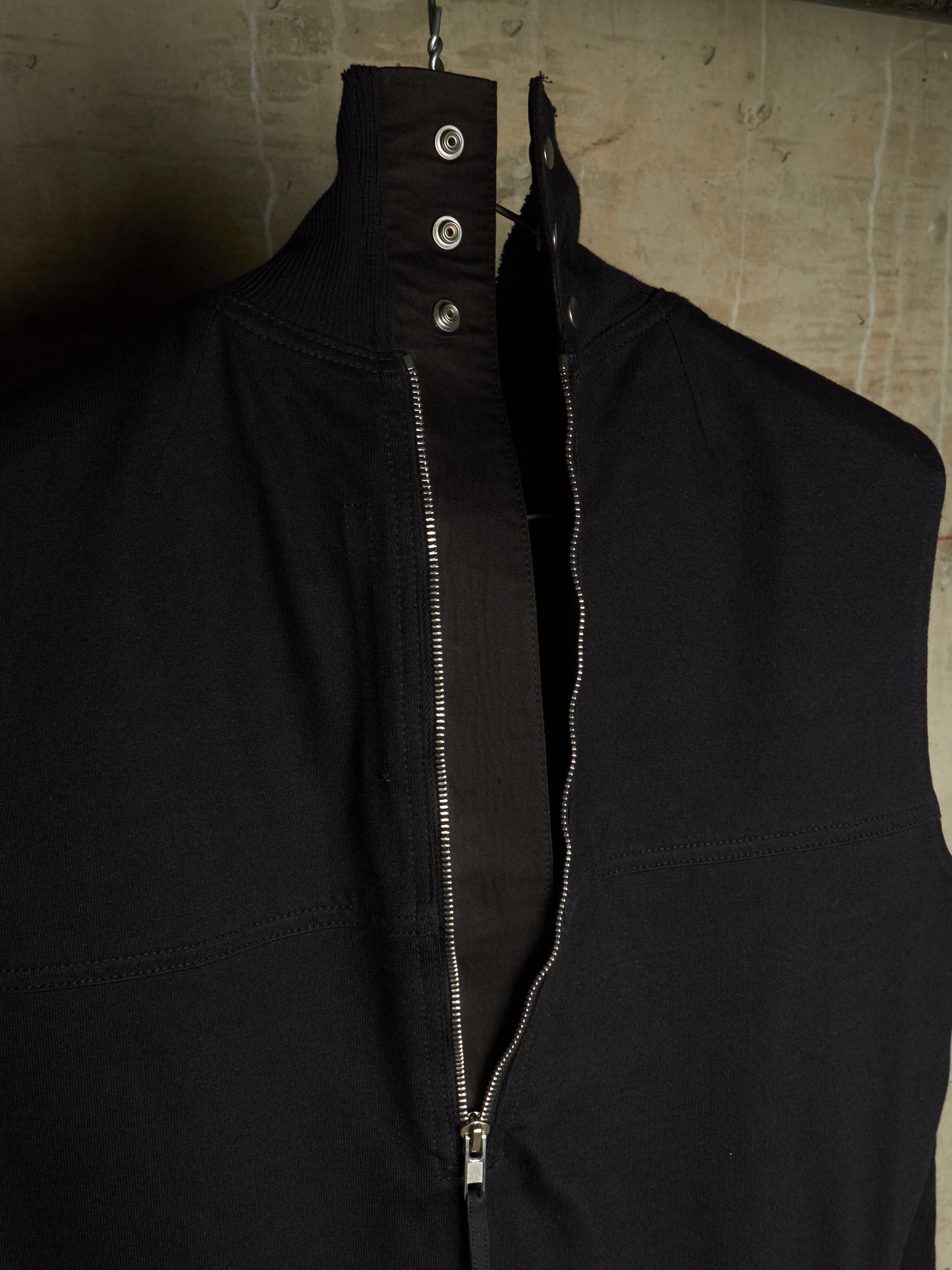 A black sleeveless zip-front vest or jacket with a high collar and three metal snap buttons at the neck, hanging against a textured concrete wall.