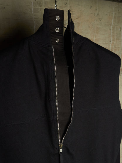 A black sleeveless zip-front vest or jacket with a high collar and three metal snap buttons at the neck, hanging against a textured concrete wall.