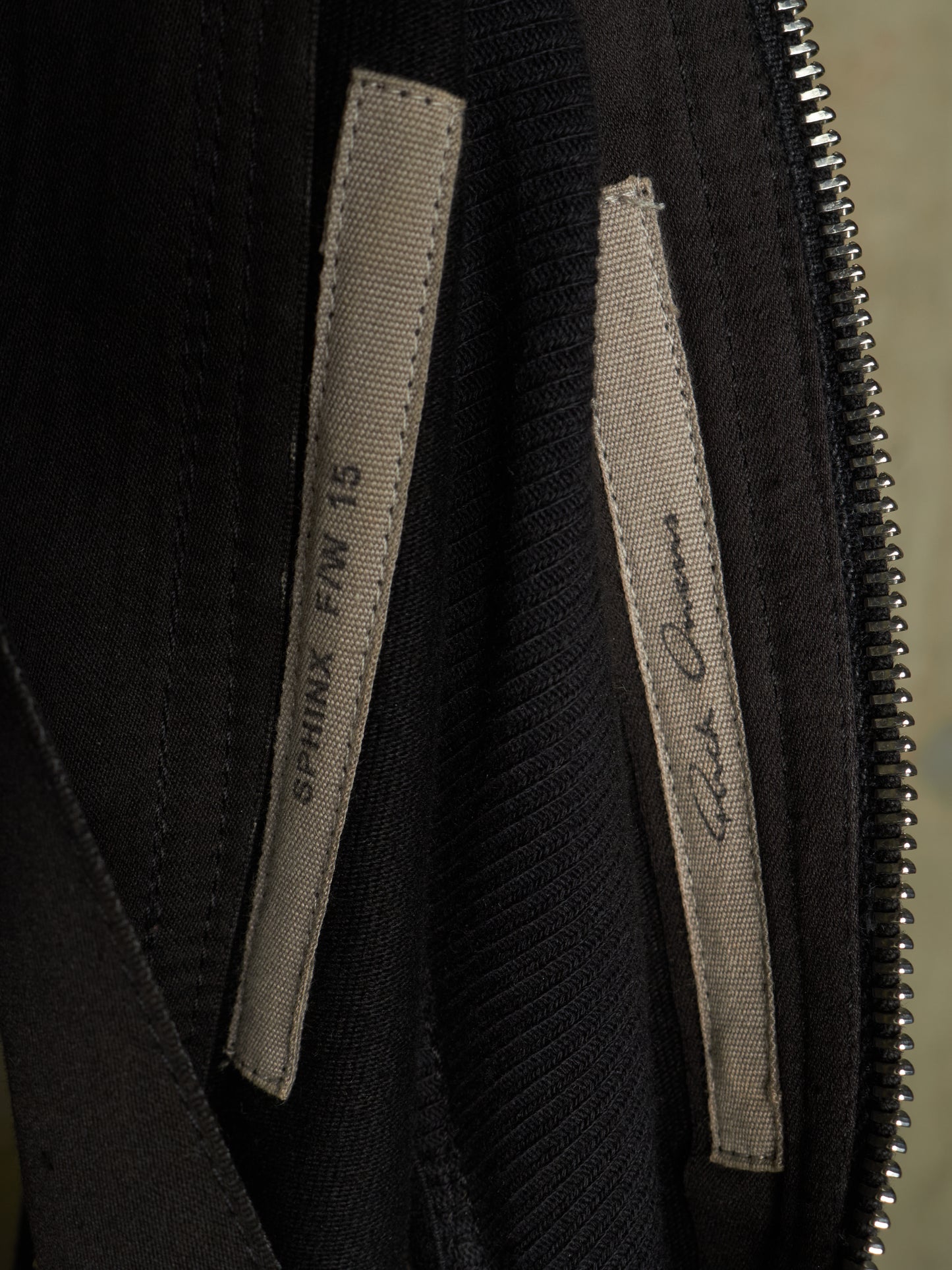 Close-up of a black garment's interior zipper and two fabric labels, one reading 'SPHINX F/W 16' and the other with a cursive signature.