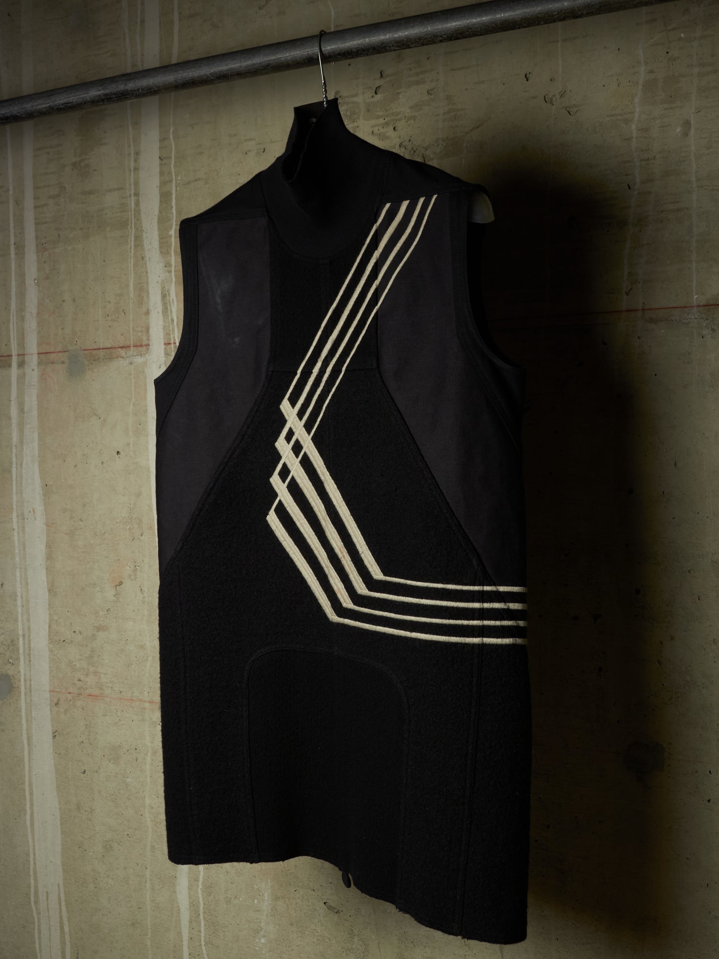 A sleeveless black dress with geometric cream-colored stripe patterns hanging on a hanger against a textured concrete wall.