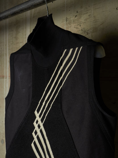 A sleeveless black vest with a high mock neck collar, featuring diagonal cream-colored knit stripes on the front, hanging on a hanger against a textured concrete wall.