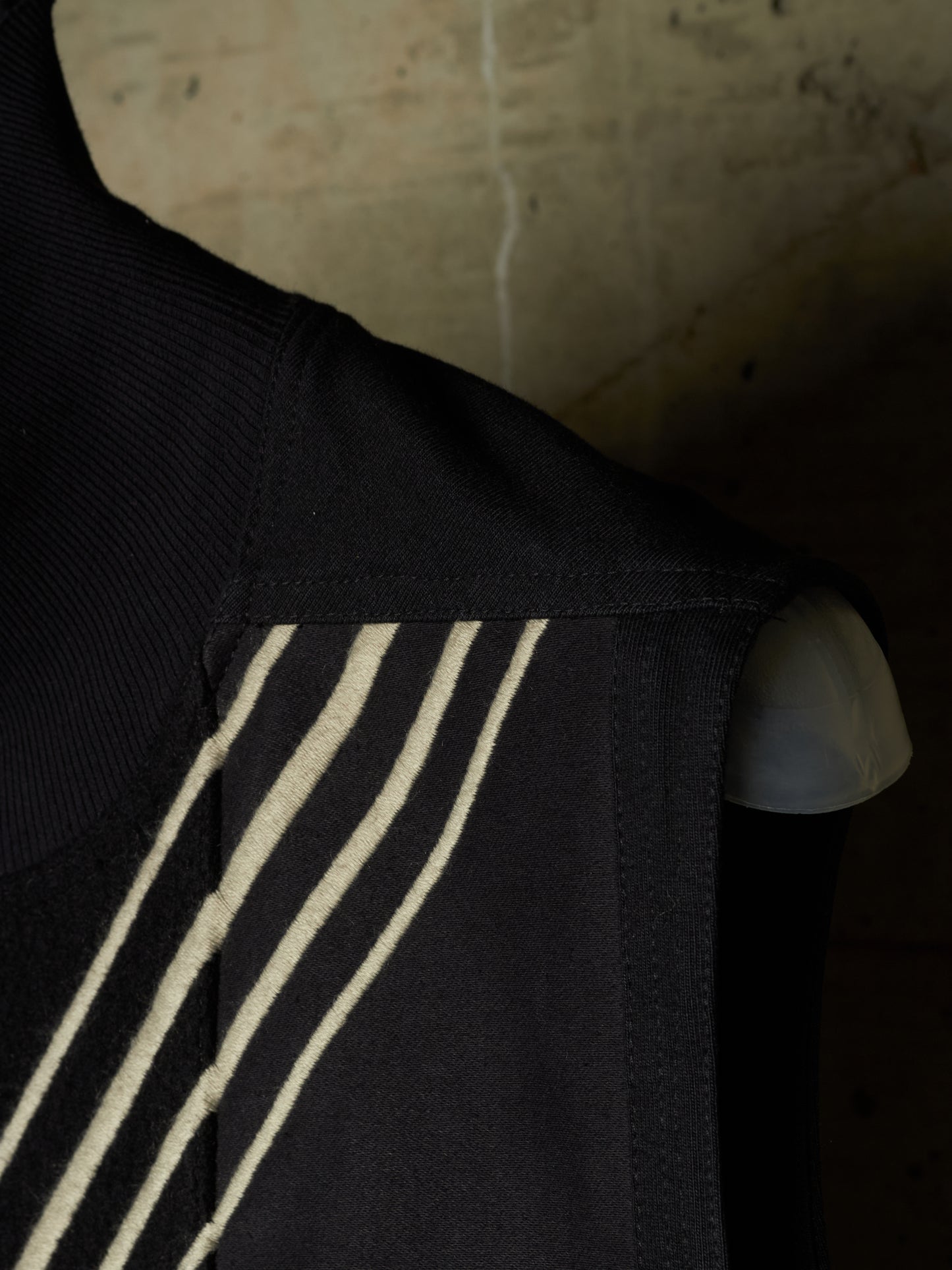 Close-up of a black sleeveless garment with three diagonal cream stripes on the shoulder, shown against a dark textured background.