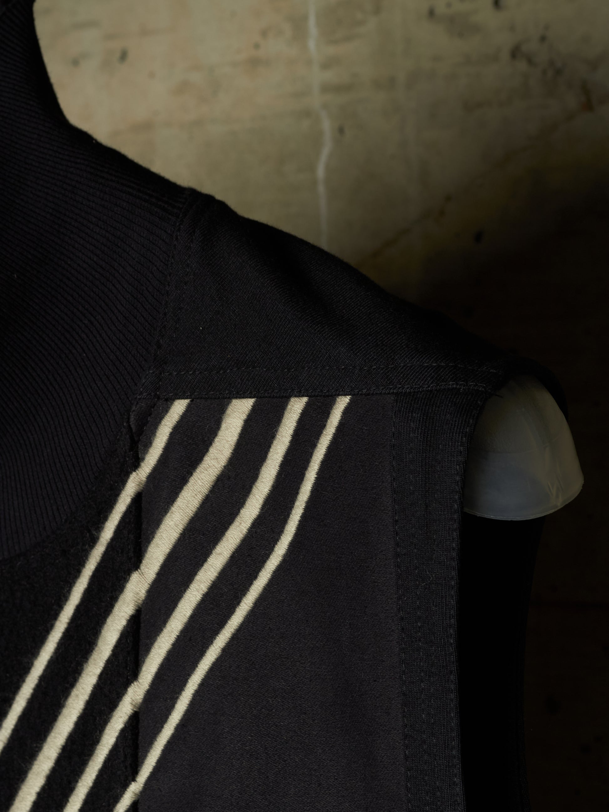 Close-up of a black sleeveless garment with three diagonal cream stripes on the shoulder, shown against a dark textured background.