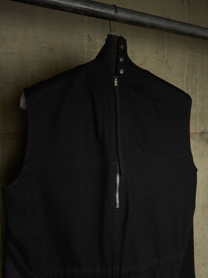 A black sleeveless zip-front vest with a high collar and snap details, hanging on a metal rod against a textured concrete wall.
