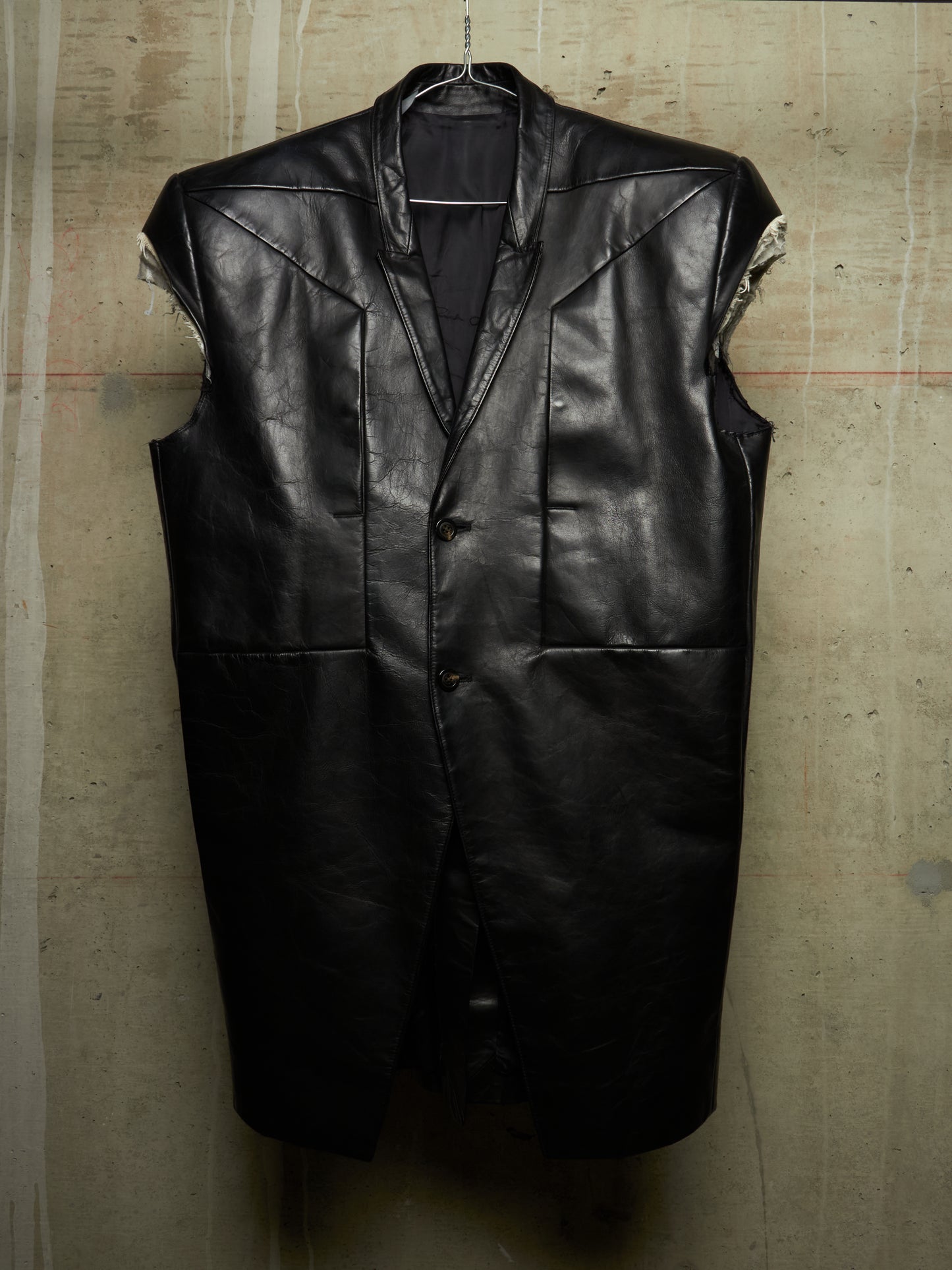 A black leather sleeveless vest with a button-front closure and structured shoulders, hanging against a textured concrete wall.