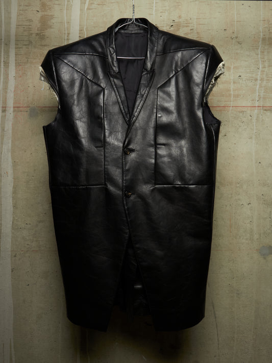 A black leather sleeveless vest with a button-front closure and structured shoulders, hanging against a textured concrete wall.