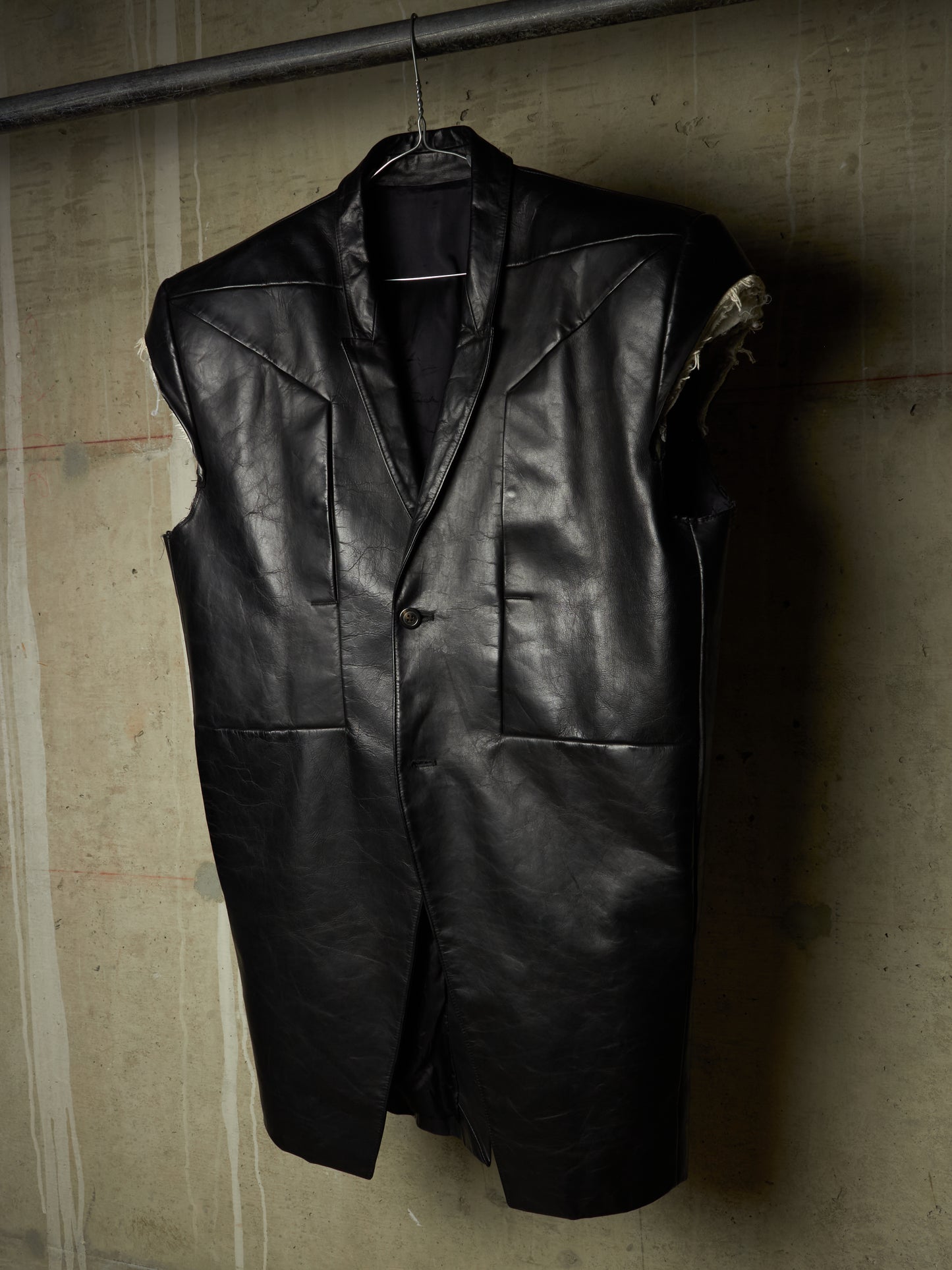 A black leather vest hanging on a hanger against a textured concrete wall.