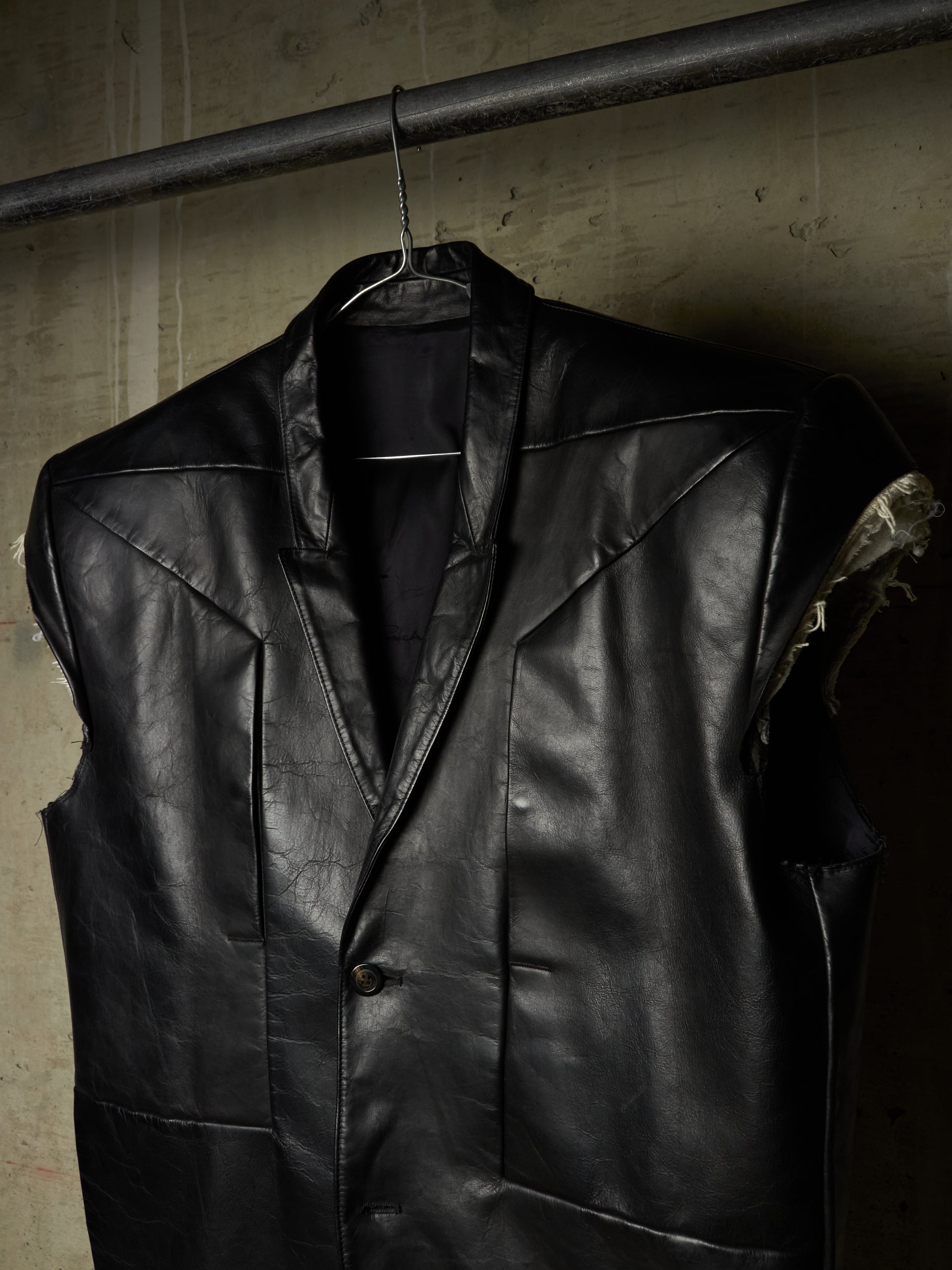 A black leather vest with a shawl collar and single front button, hanging on a metal hanger from a dark industrial rod against a textured concrete wall.
