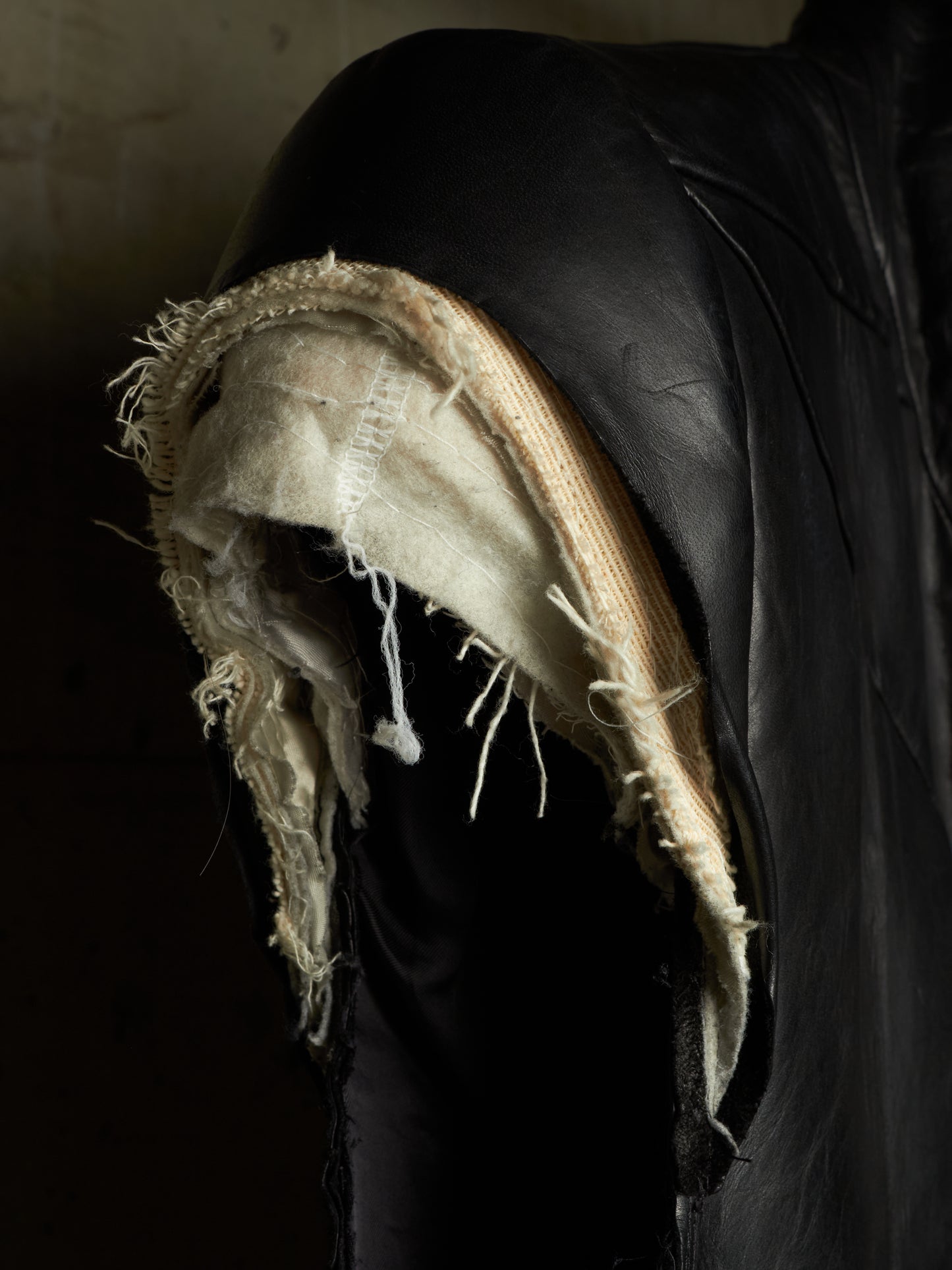 A close-up of a black leather garment, likely a jacket or coat, showing a heavily distressed and frayed tear on the shoulder with off-white and beige fabric layers exposed.