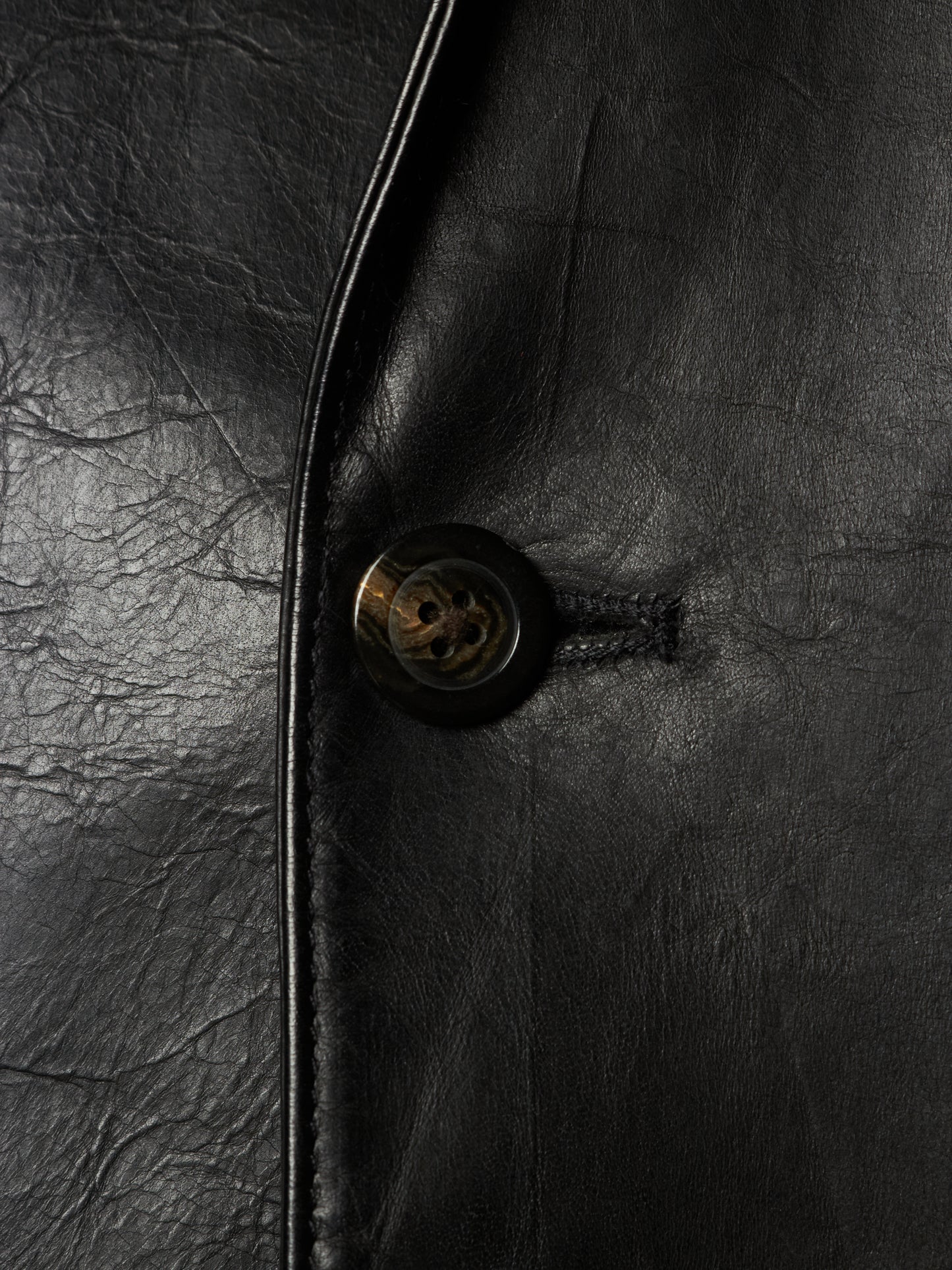 Close-up of a black leather jacket showing the texture of the leather, a black button with a gold-toned center, and a dark metal zipper track.