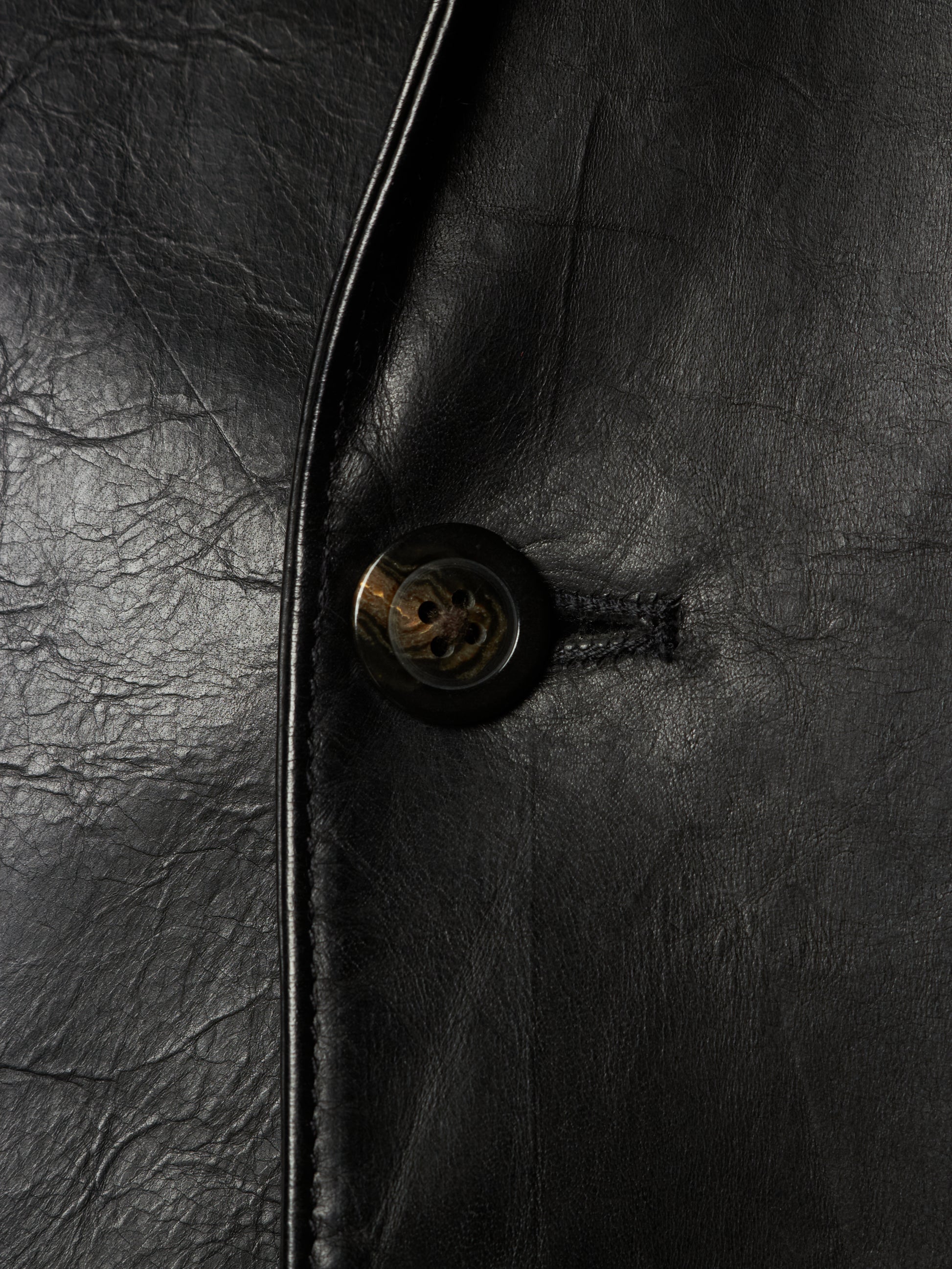 Close-up of a black leather jacket showing the texture of the leather, a black button with a gold-toned center, and a dark metal zipper track.