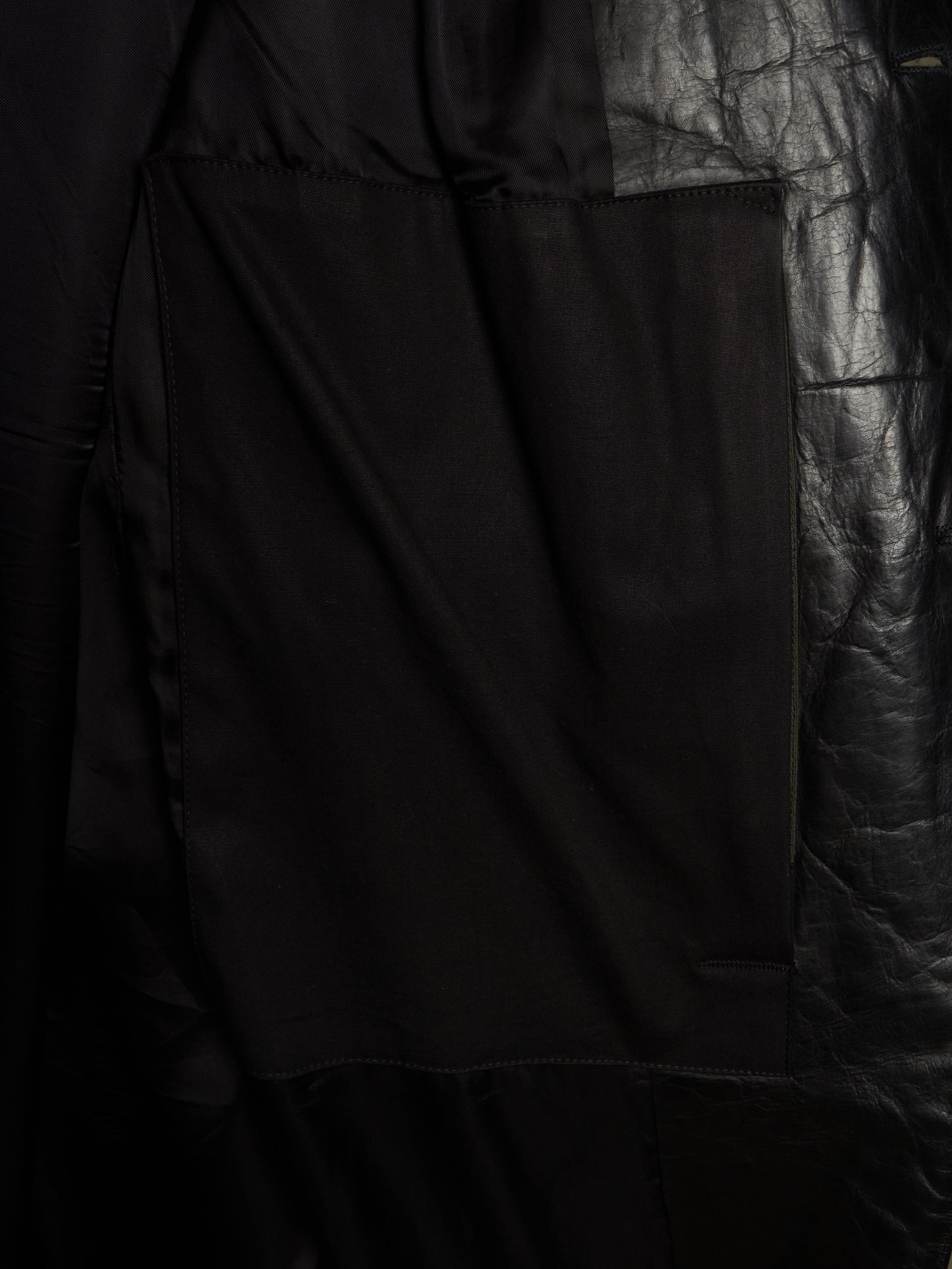 Close-up detail of a black garment showing a large rectangular patch pocket with a textured, slightly ribbed fabric body and a crinkled black leather or faux-leather panel on the right side.