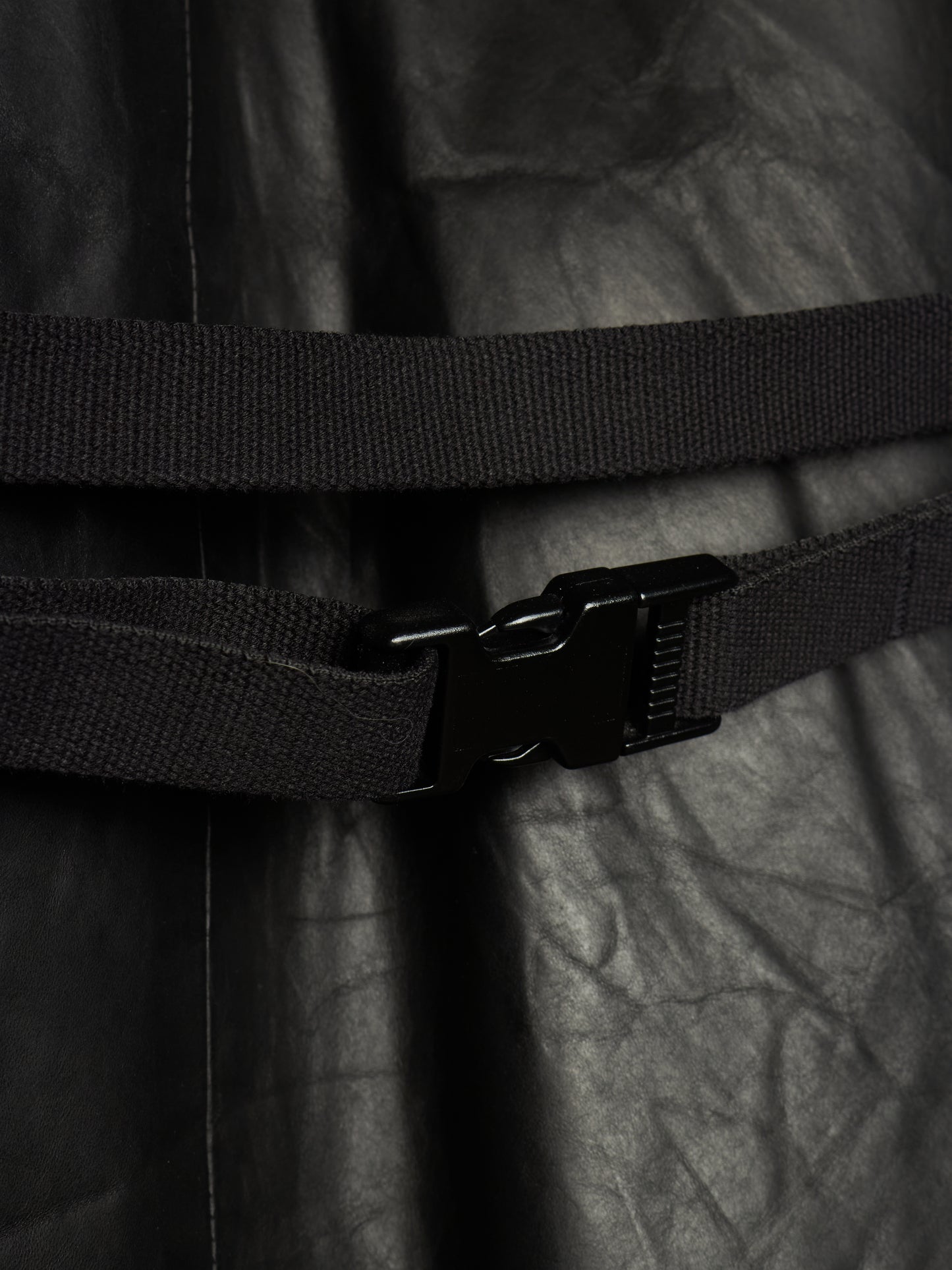 Close-up of a black adjustable web belt with a black plastic quick-release buckle, wrapped around a black crinkled fabric garment.