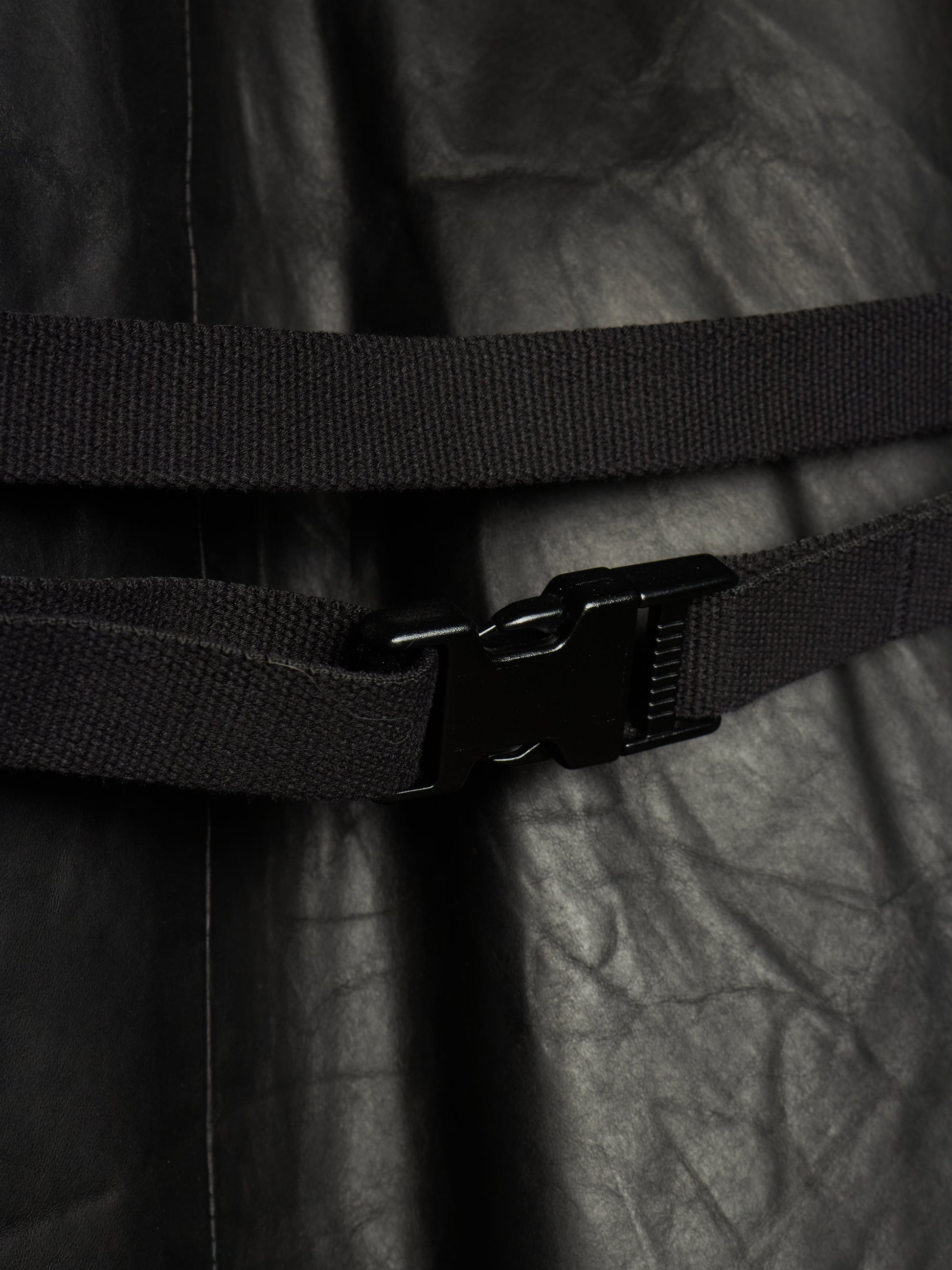 Close-up of a black adjustable web belt with a black plastic quick-release buckle, wrapped around a black crinkled fabric garment.