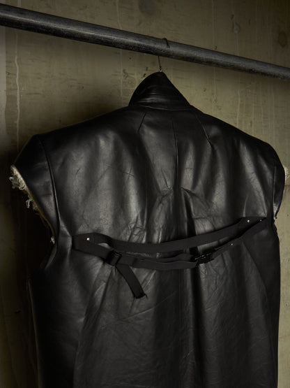 A black leather sleeveless vest or cape-like garment hanging on a metal rod against a textured concrete wall, with a black strap detail across the back.