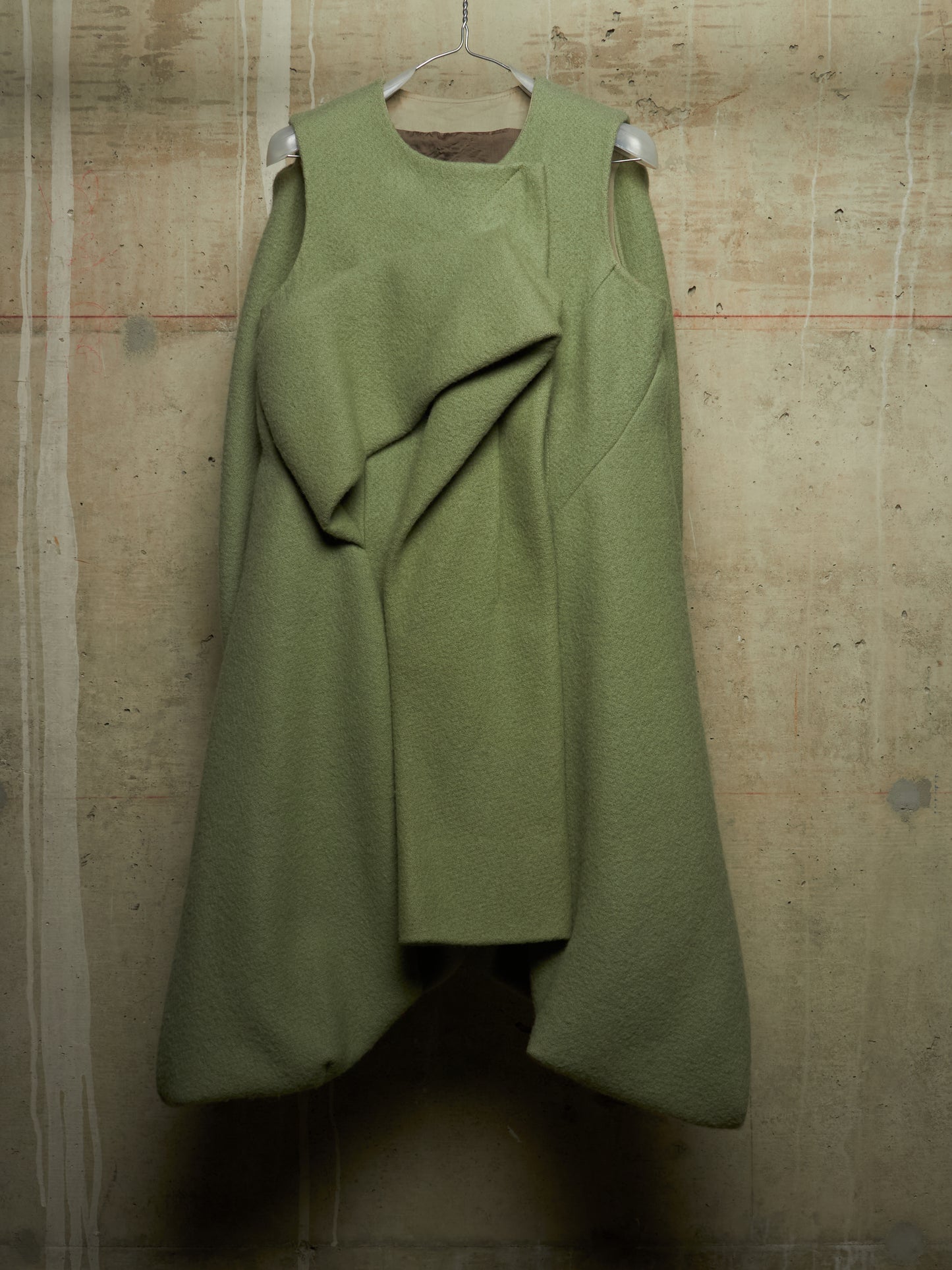 An olive green sleeveless dress with a draped, asymmetrical front detail, hanging on a wire hanger against a textured concrete wall.