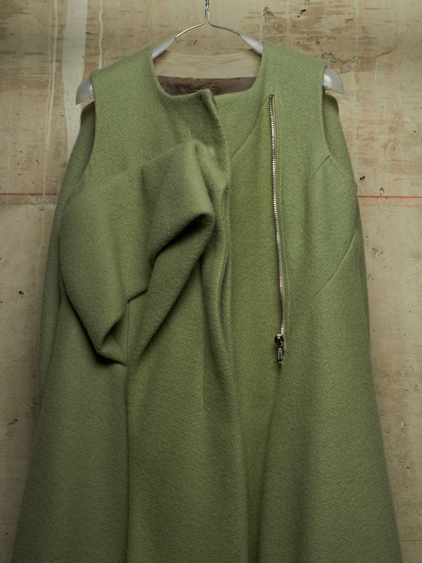 An olive green sleeveless wool coat with a draped front detail and a vertical silver zipper, hanging on a metal hanger against a textured concrete wall.