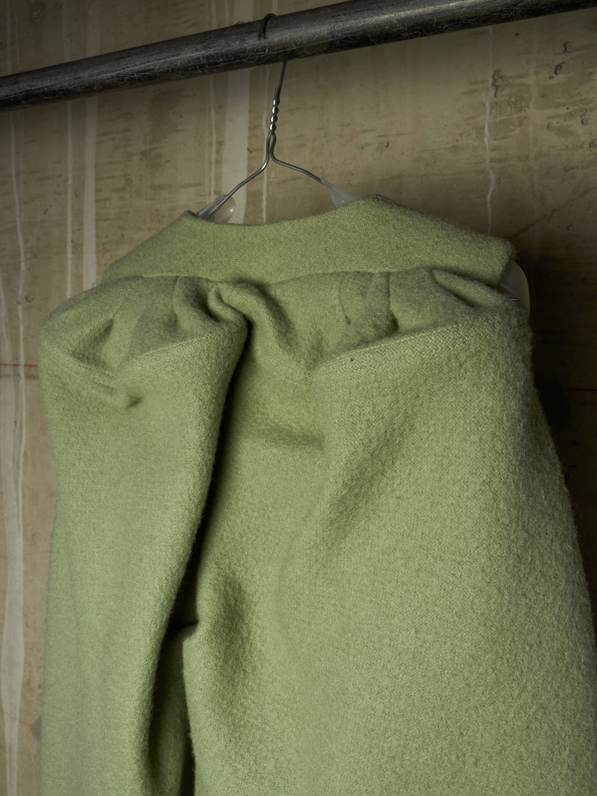 A sage green wool coat draped over a metal hanger, hanging from a dark metal clothing rack against a textured, warm-toned wall.