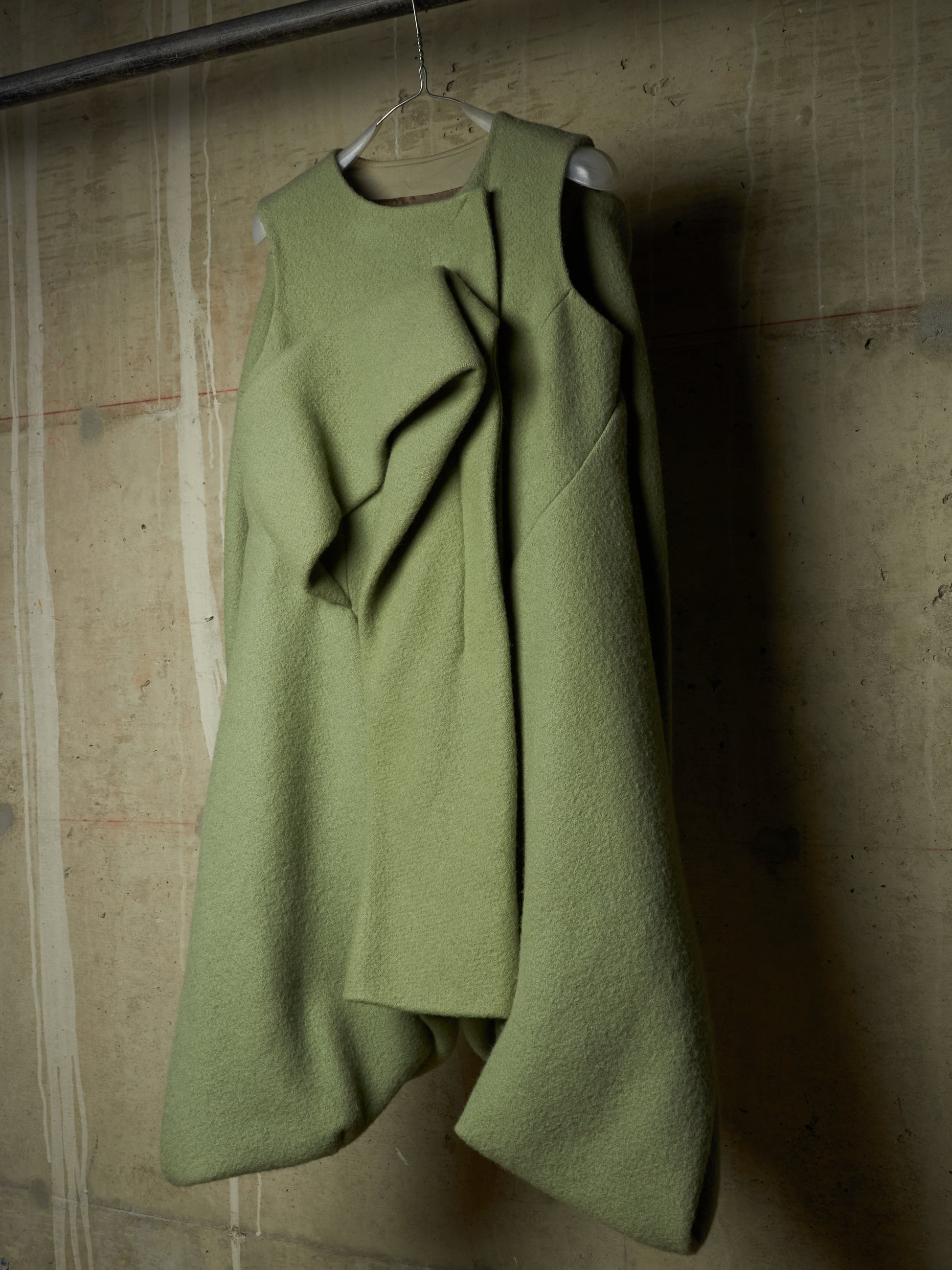 An asymmetrical olive green wool coat with draped ruffle details, hanging on a wire hanger against a textured concrete wall.