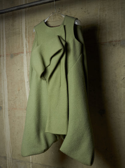 An asymmetrical olive green wool coat with draped ruffle details, hanging on a wire hanger against a textured concrete wall.