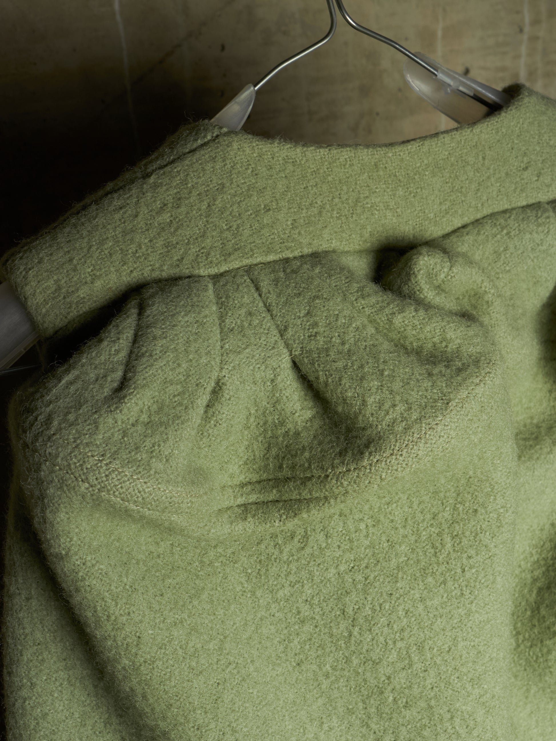 A close-up of a textured sage green knit garment draped on a metal hanger against a dark, neutral background.