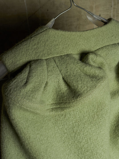 A close-up of a textured sage green knit garment draped on a metal hanger against a dark, neutral background.