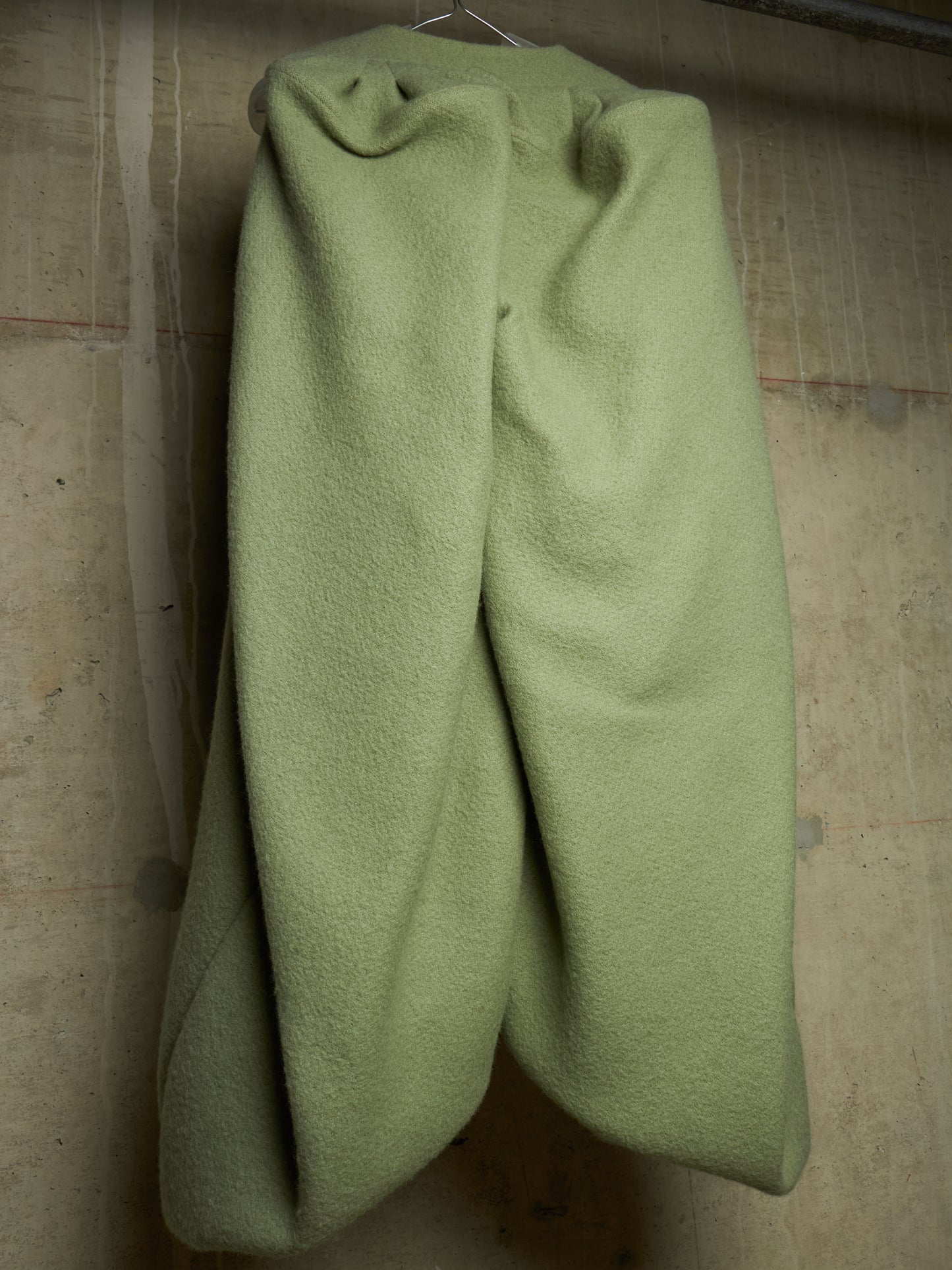 A pair of light sage green wool trousers hanging on a metal hanger against a textured concrete wall.