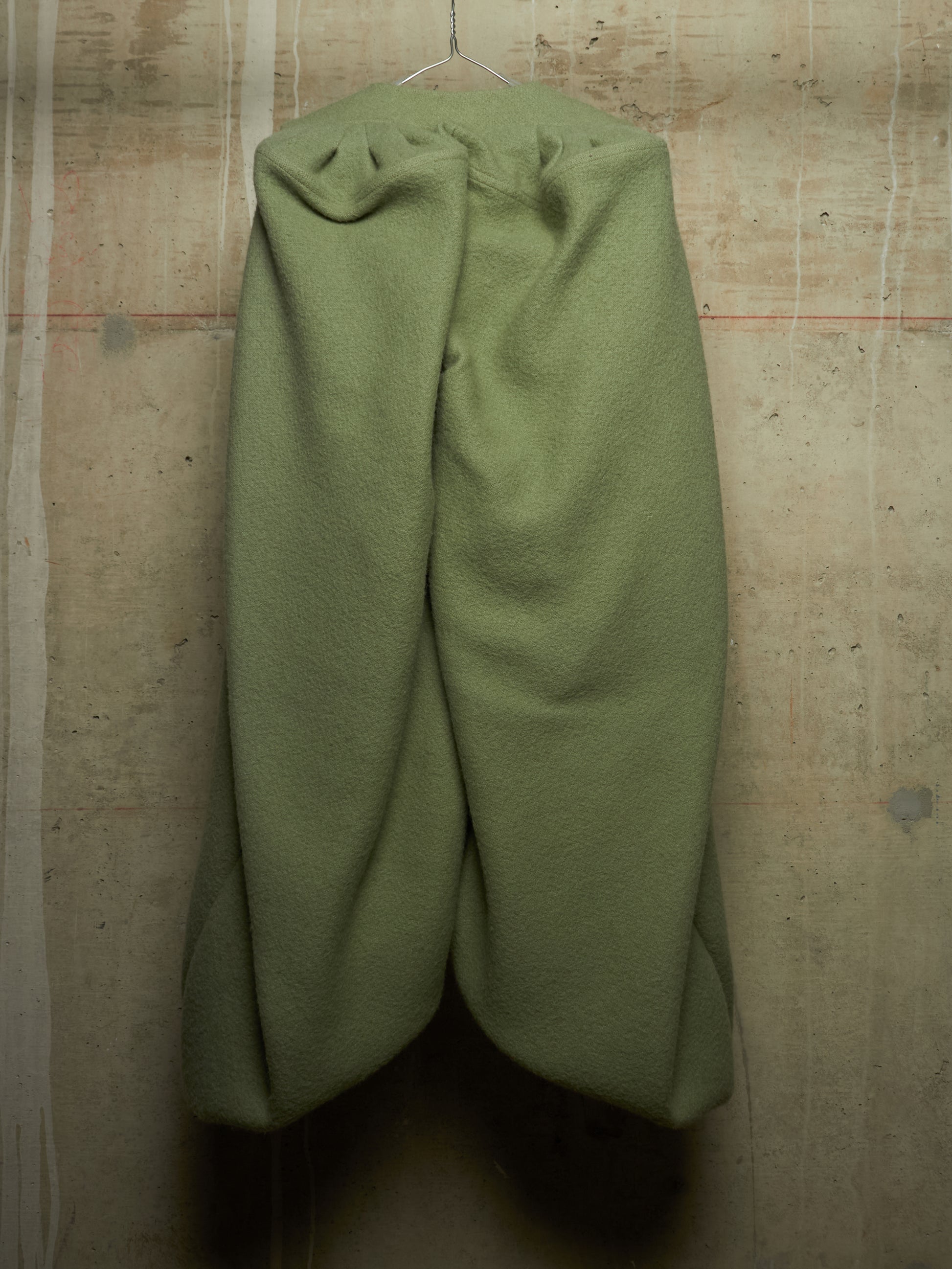 Olive green wool trousers hanging from a metal hanger against a textured concrete wall.